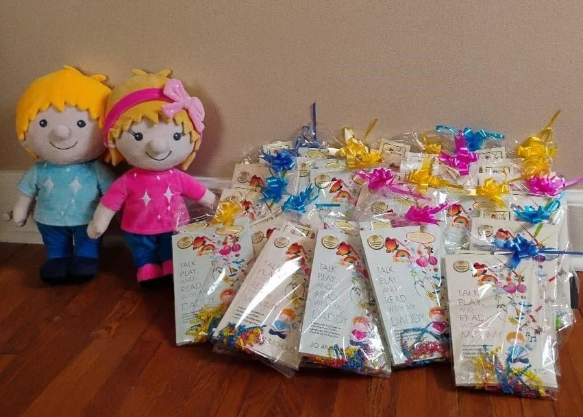 Two large plush toys, a boy and a girl, stand next to a pile of brightly decorated gift bags on a wooden floor against a beige wall. The boy plush has blonde hair, a blue shirt, and dark pants. The girl plush has blonde hair with a pink headband and bow, a pink shirt, and pink and dark pants.