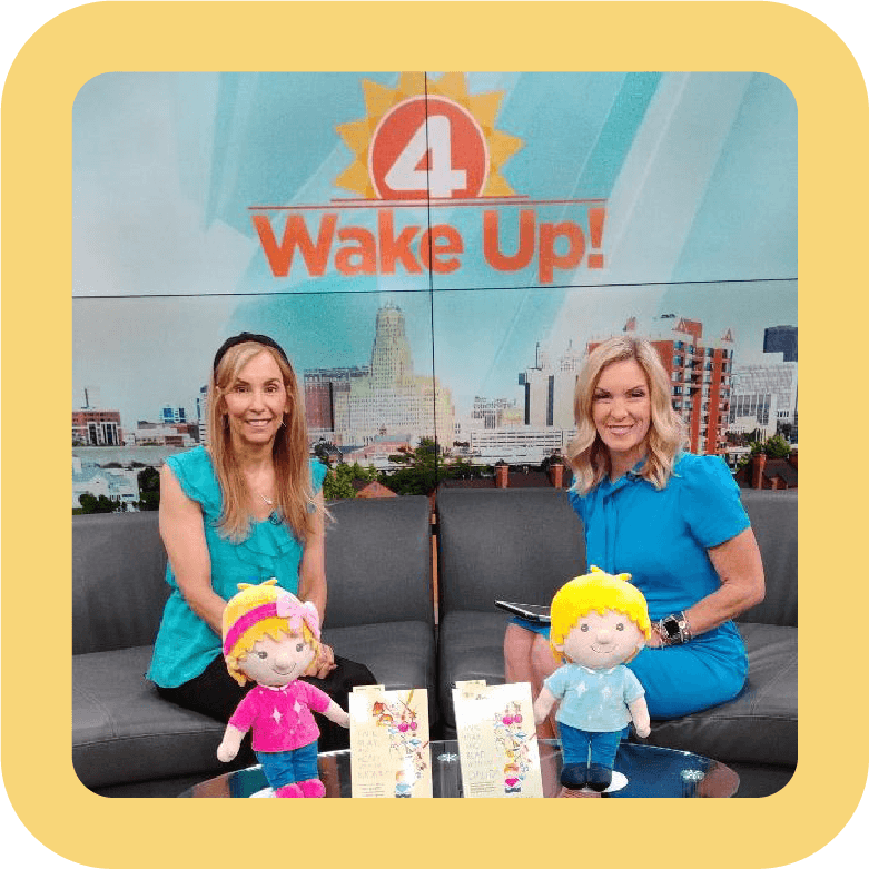 Two women sitting on a TV set with a city skyline background, smiling at the camera, with two plush toys and two cards on a table in front of them. A large screen behind them displays a logo that says '4 Wake Up!'