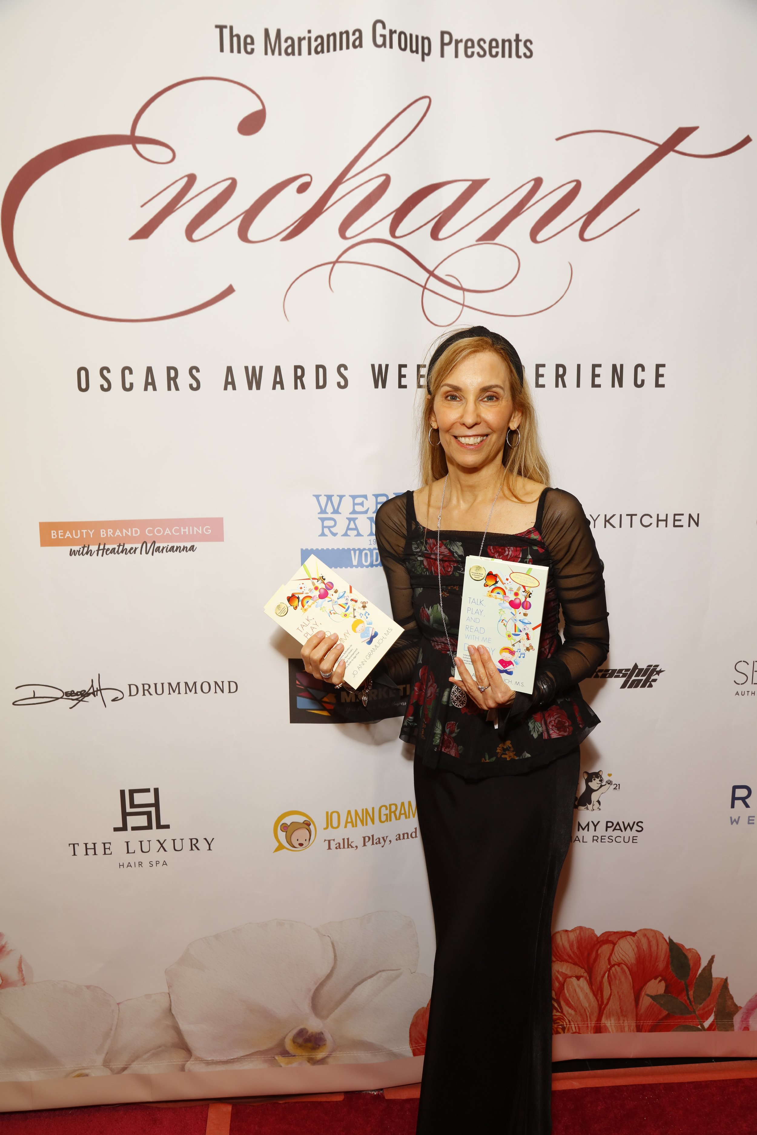 A woman, Jo Ann Gramlich, standing on a red carpet holding two pamphlets at the Enchant Oscars awards week experience event, with a backdrop featuring the event's branding and various sponsor logos.