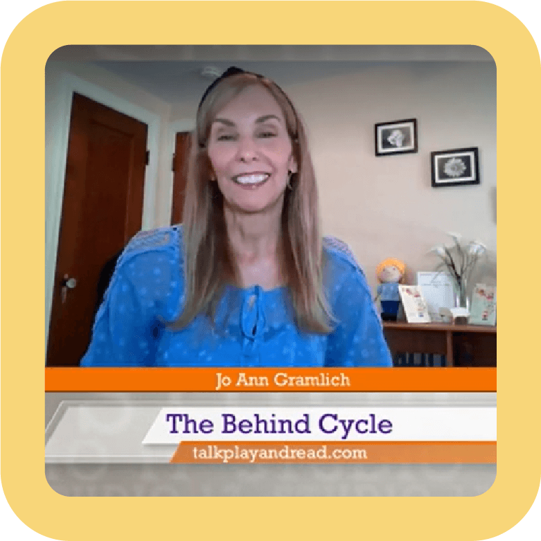 A woman with long blonde hair wearing a blue blouse, smiling in a room with beige walls and framed pictures, sitting behind a desk or table. The screen displays her name, Jo Ann Gramlic, and the title "The Behind Cycle" with a website link talkplayandread.com.