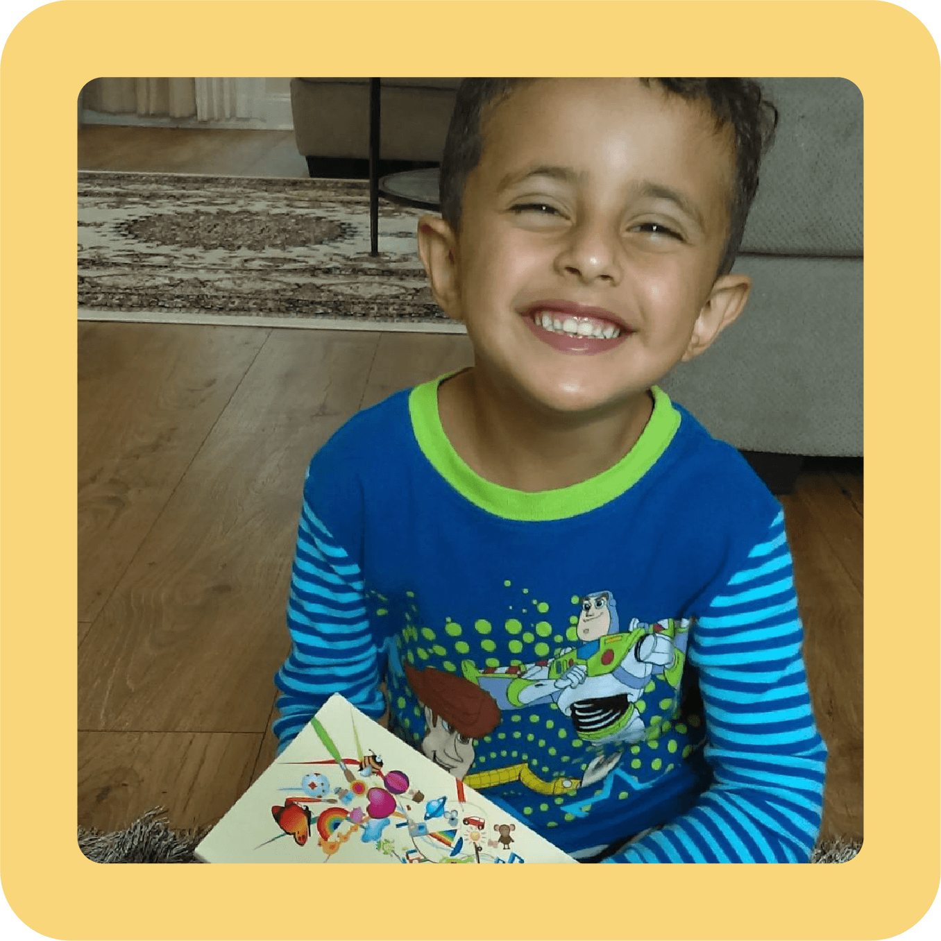 A smiling young boy with dark hair, wearing a blue and green Toy Story-themed long-sleeve shirt, sitting on a hardwood floor, holding a colorful sticker book.