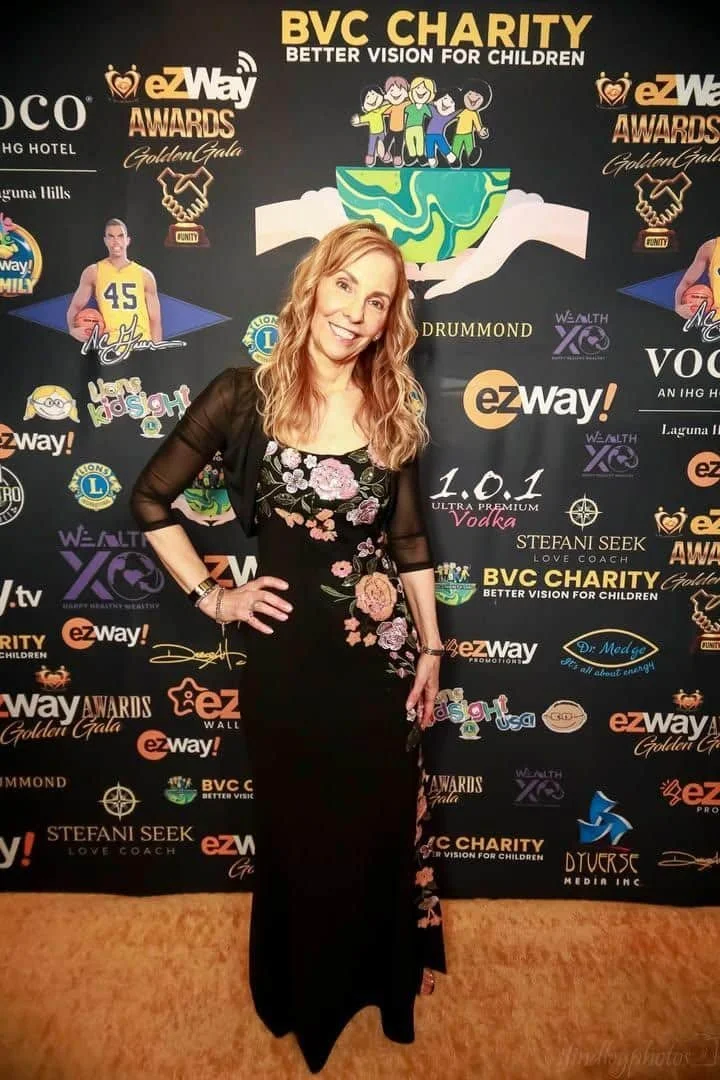 A woman, jo Ann Gramlich, in a long black floral dress and sheer black jacket standing in front of a step and repeat banner at a charity event. The banner features logos, illustrations, and text promoting BVC Charity and various sponsors.