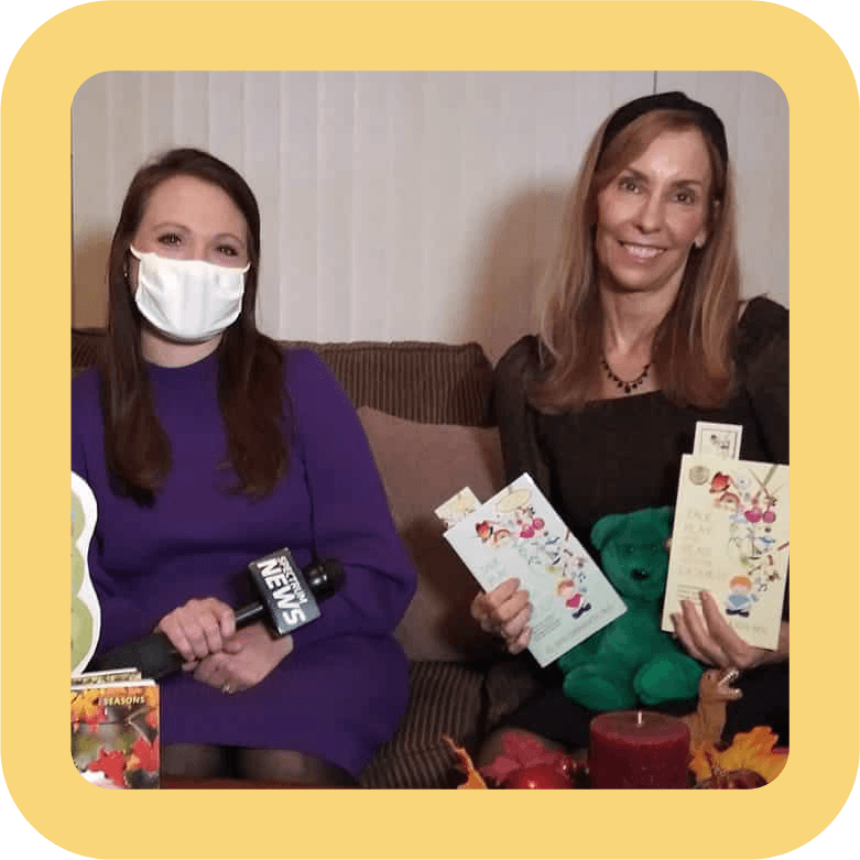 Two women sitting on a couch, one wearing a purple dress and a white face mask, holding a microphone with a news logo, and the other wearing a black top, holding books, with a pillow and decorations on the couch.
