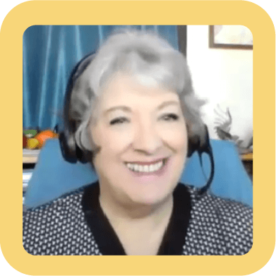 Smiling elderly woman with gray hair wearing a headset, sitting in front of a blue curtain with a pattern and a desk.