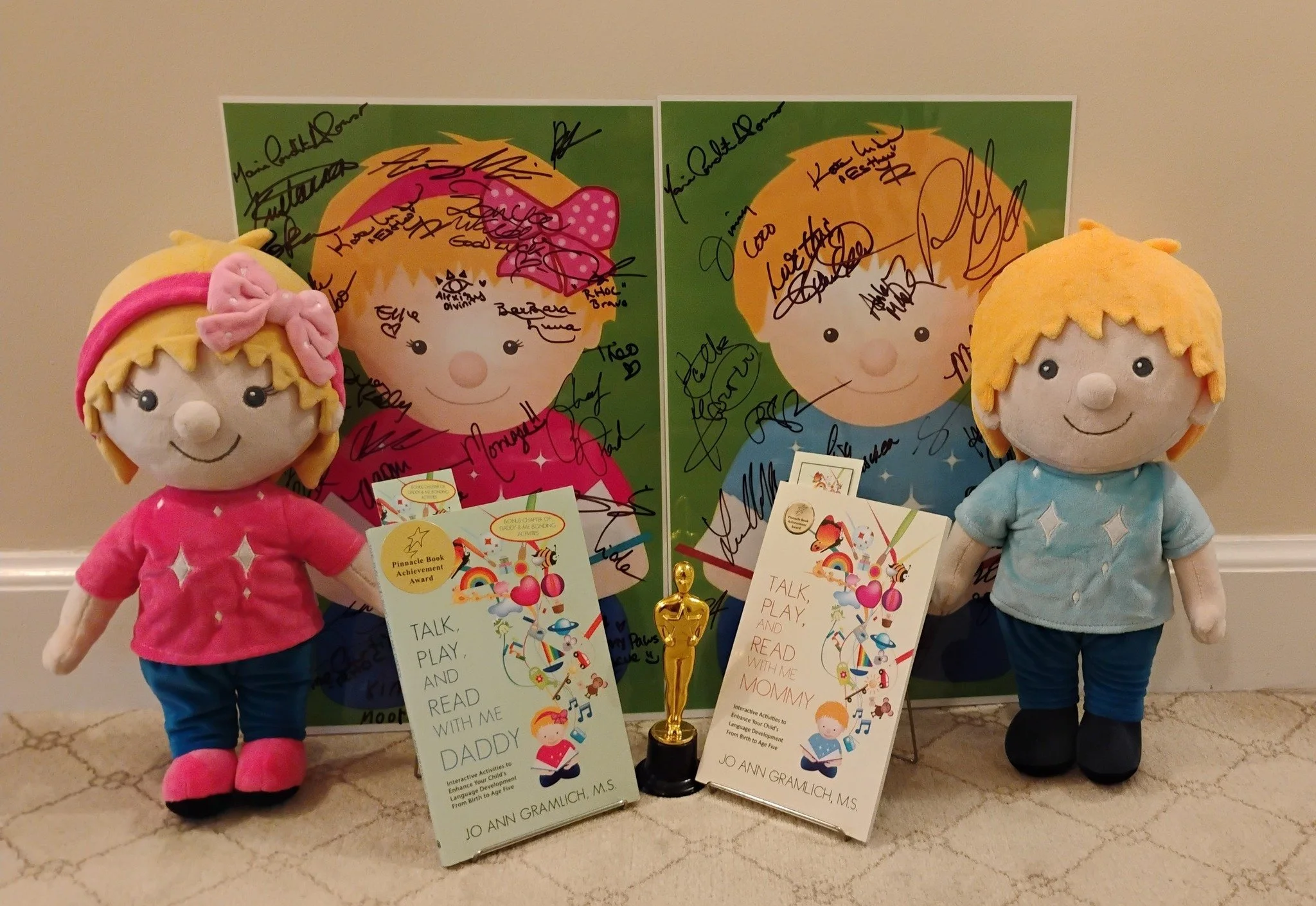 Two plush dolls of children with signed posters and a small award trophy on a beige carpet. One doll has blonde hair with a pink headband, wearing a pink shirt and blue pants. The other has blonde hair, wearing a light blue shirt and dark blue pants.