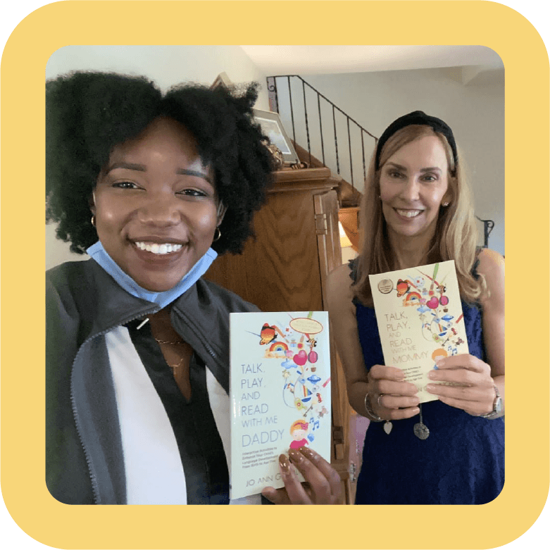 Two women smiling and holding books titled "Talk, Play, and Read with Me Daddy" and "Talk, Play, and Read with Me Mommy" inside a home.