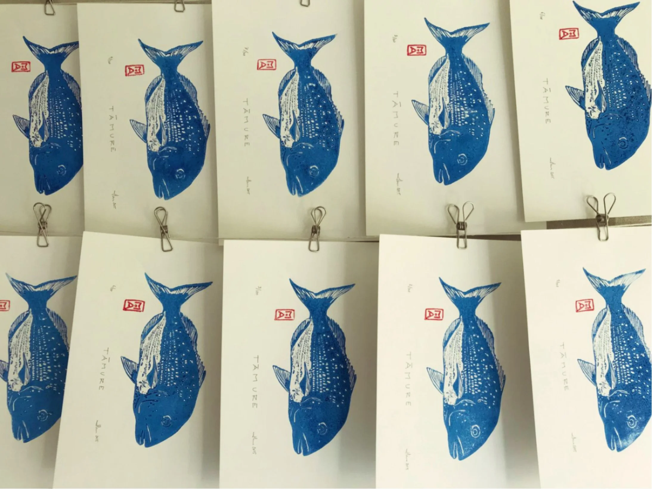 Multiple white cards with a blue fish illustration and the words 'TAMURE' and a signature, attached to a wire with small clips.