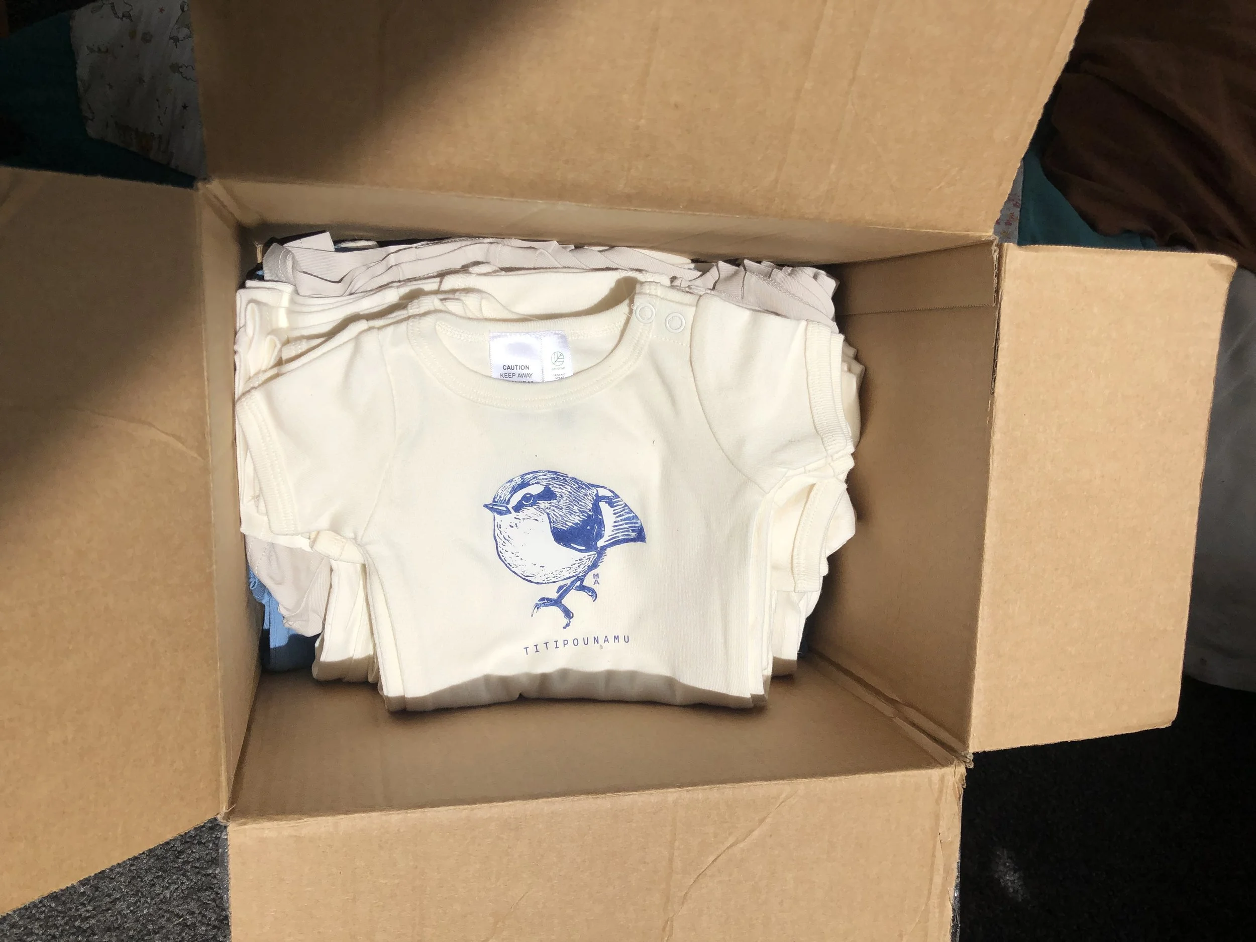 A cardboard box containing folded baby T-shirts, including one with a blue bird print and the text 'TITIPOUNAMU'.