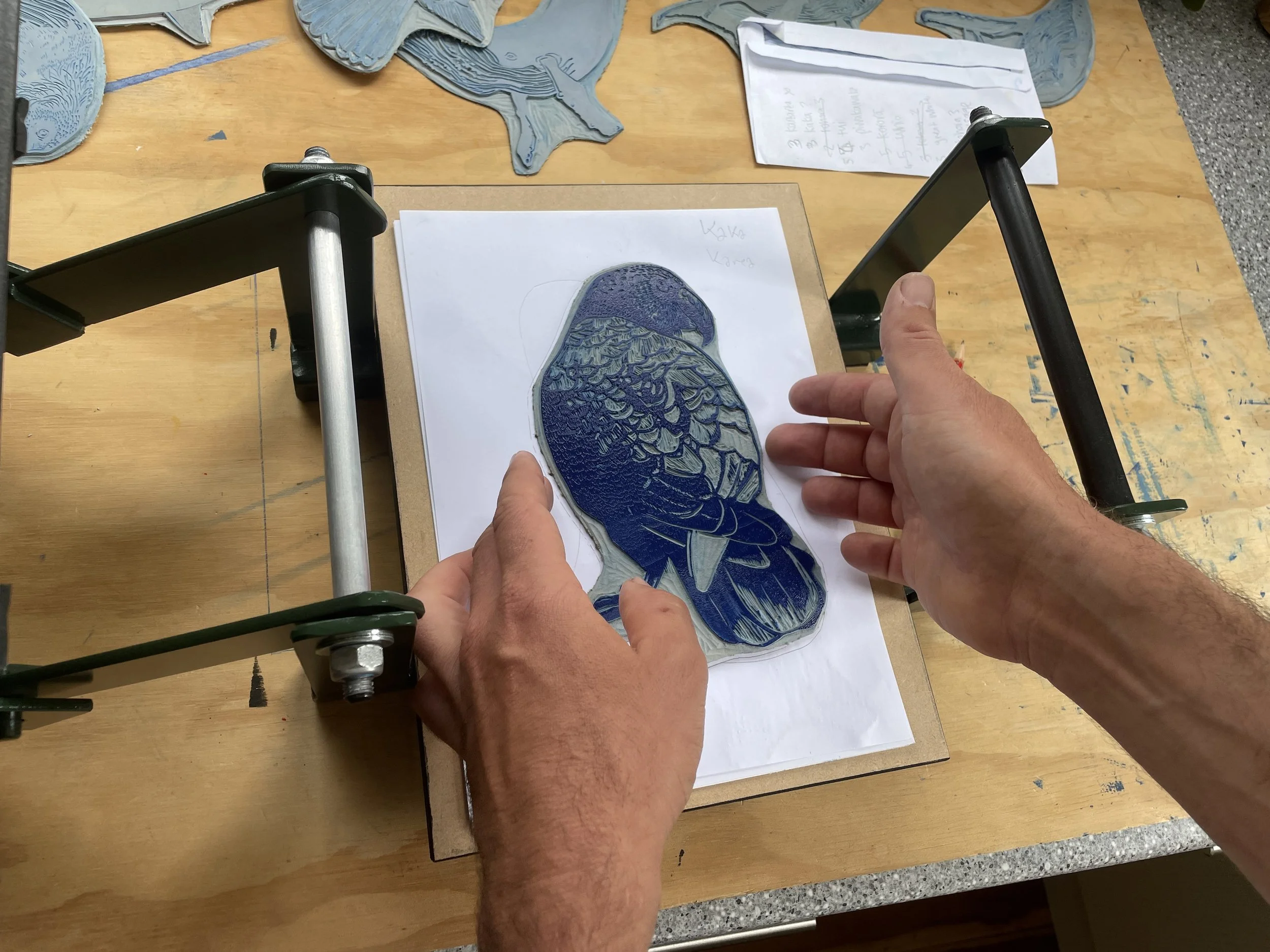 Person handling a blue embroidered patch of a bird on a wooden table with pattern paper, gloves in the background, and handwritten notes on a paper.