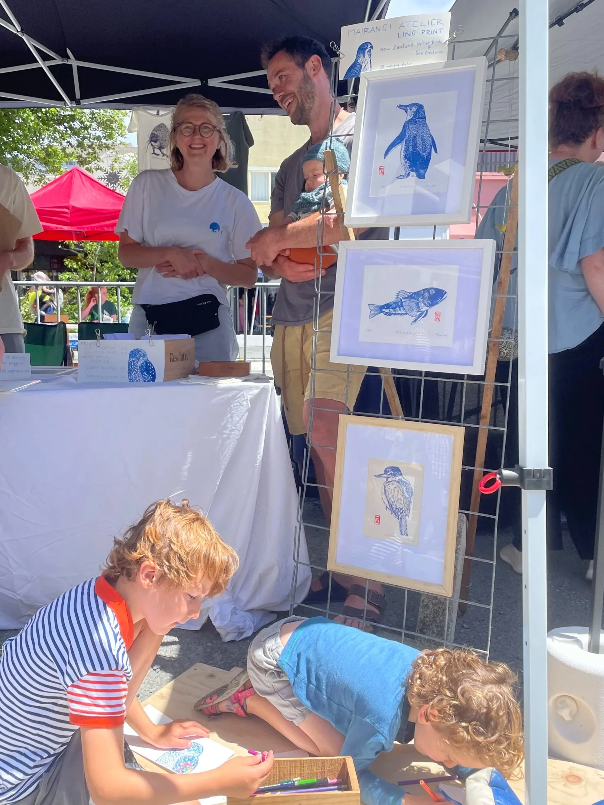Two adults, a man and a woman, at an art booth with framed bird-themed artwork, while two children are drawing on a ground board in front of the booth under sunlight.