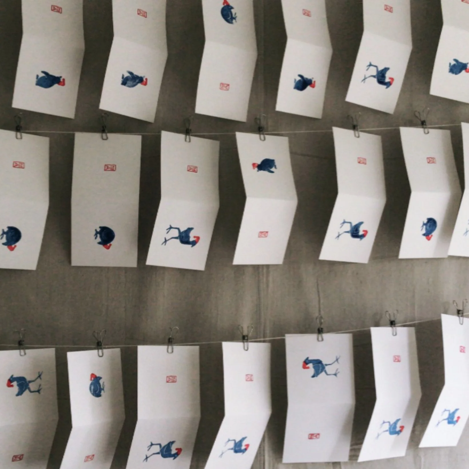 Multiple sheets of paper hanging on lines with photographs of a cartoon blue ostrich wearing a red hat, falling or upside-down, attached with clips.