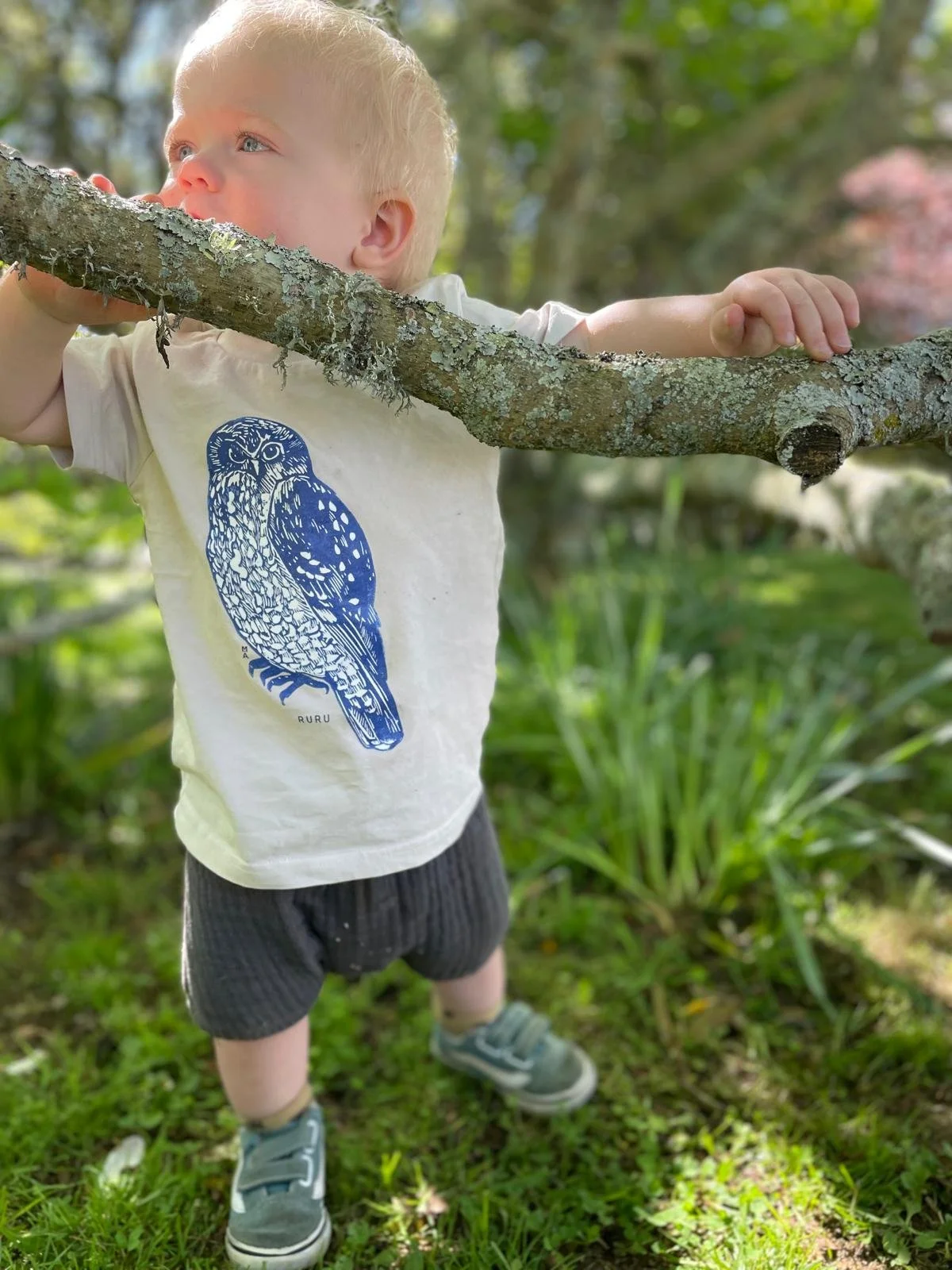 A young boy with blonde hair is standing outdoors in a green, grassy area, holding onto a large tree branch with both hands, looking to the side with a thoughtful expression.