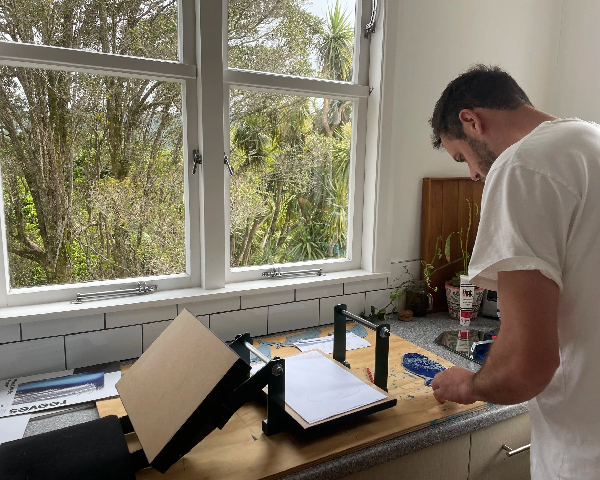 A man working at a wooden desk with an adjustable printing press, in a kitchen with large windows overlooking trees and greenery outside.