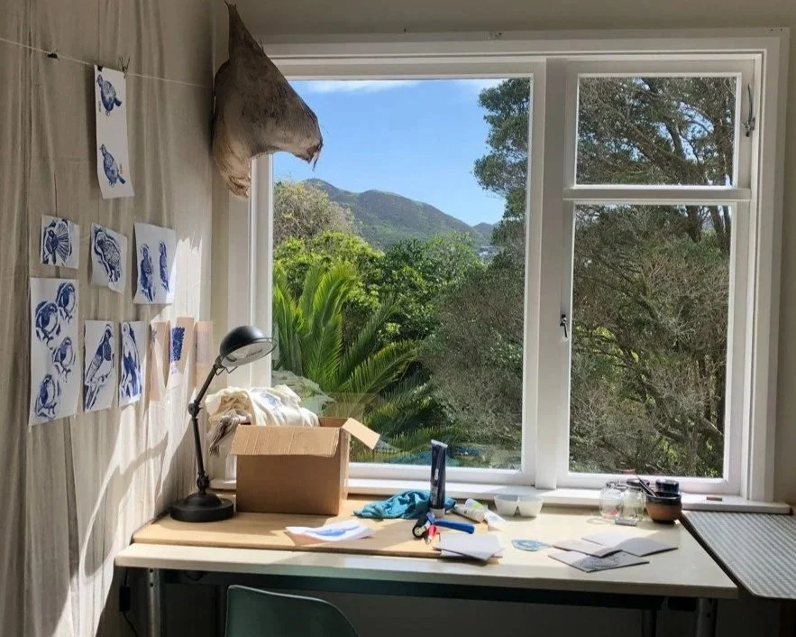 A cluttered desk next to a window with a view of trees and mountains outside. The desk has papers, a lamp, a cardboard box, and some glass jars. There are blue and white illustrations of animals hanging on the wall.