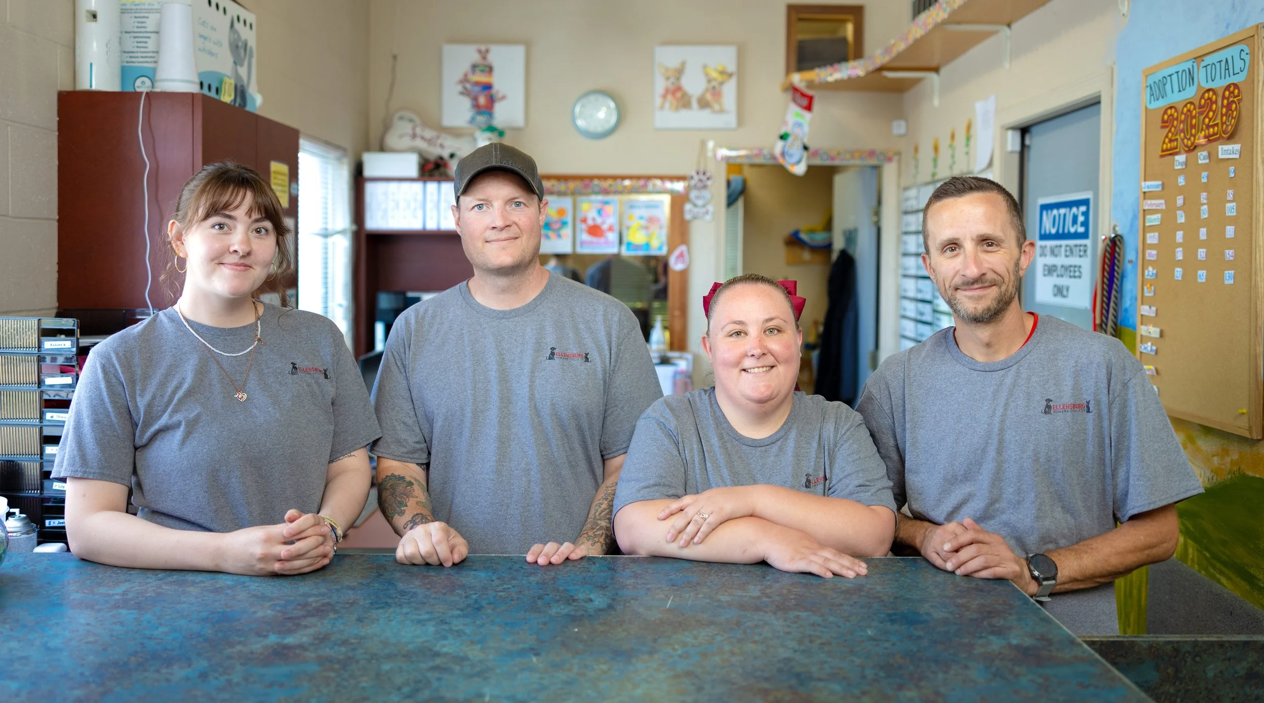 Four staff members standing behind a counter in a classroom or office environment, wearing matching gray T-shirts with a logo. They are smiling and looking at the camera, with colorful decorations and posters on the walls in the background.
