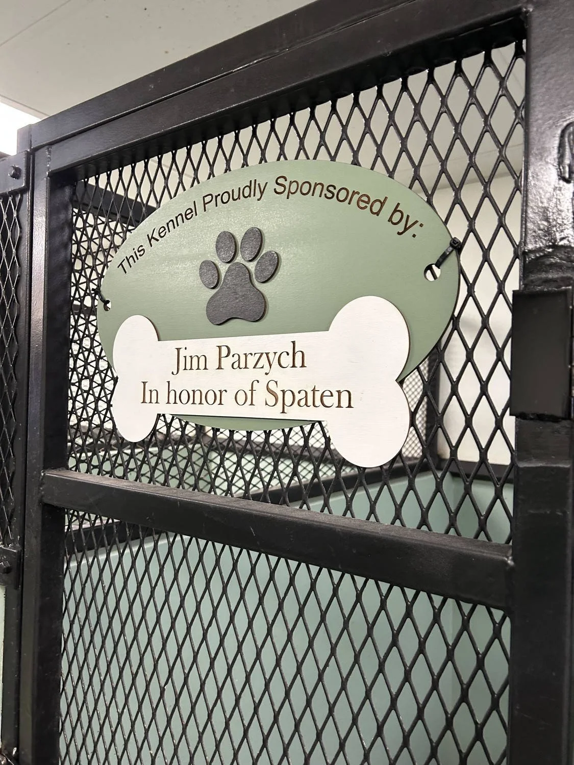 A sign on a black metal fence reading, 'This Kennel Proudly Sponsored by: Jim Parzych In honor of Spaten', with a paw print and a bone-shaped design.