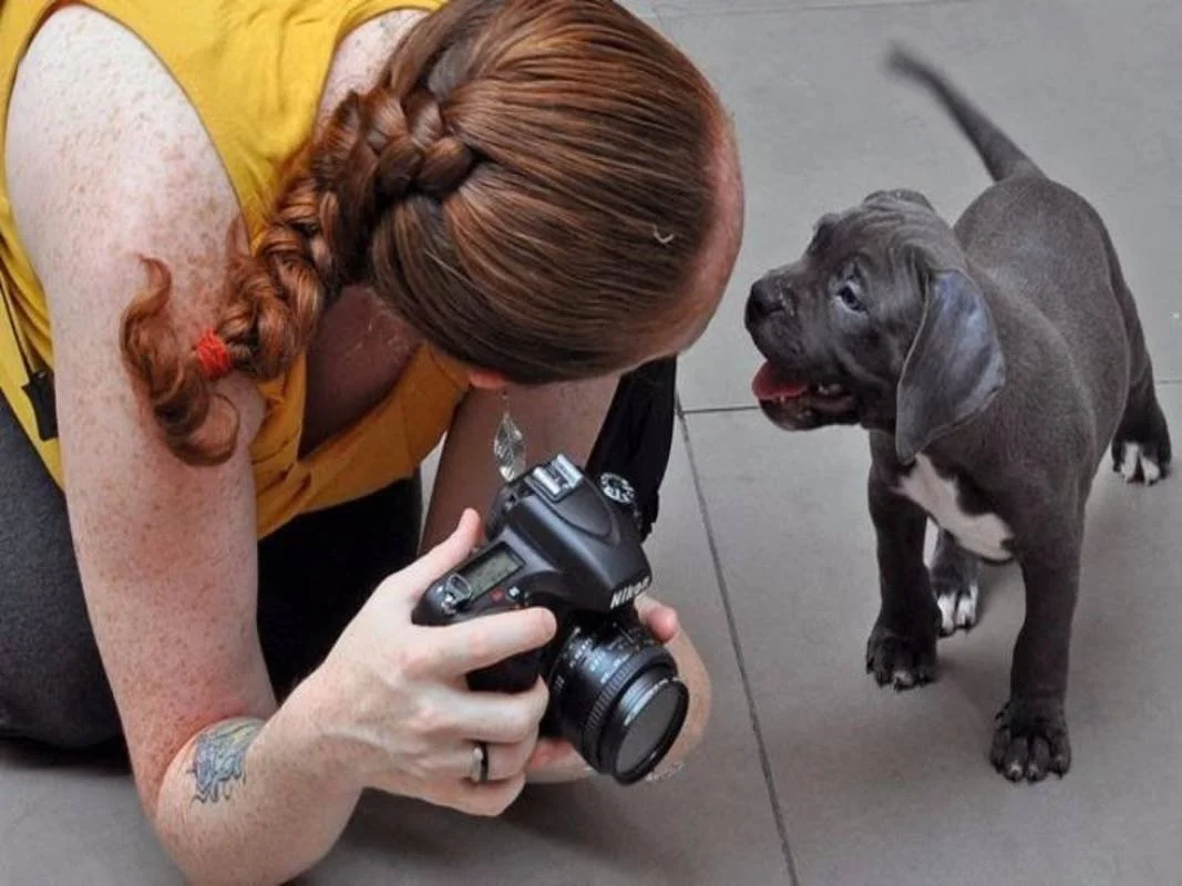 A woman with a red braid holds a camera while lying on the floor, facing a puppy. The puppy looks at her with a happy expression.