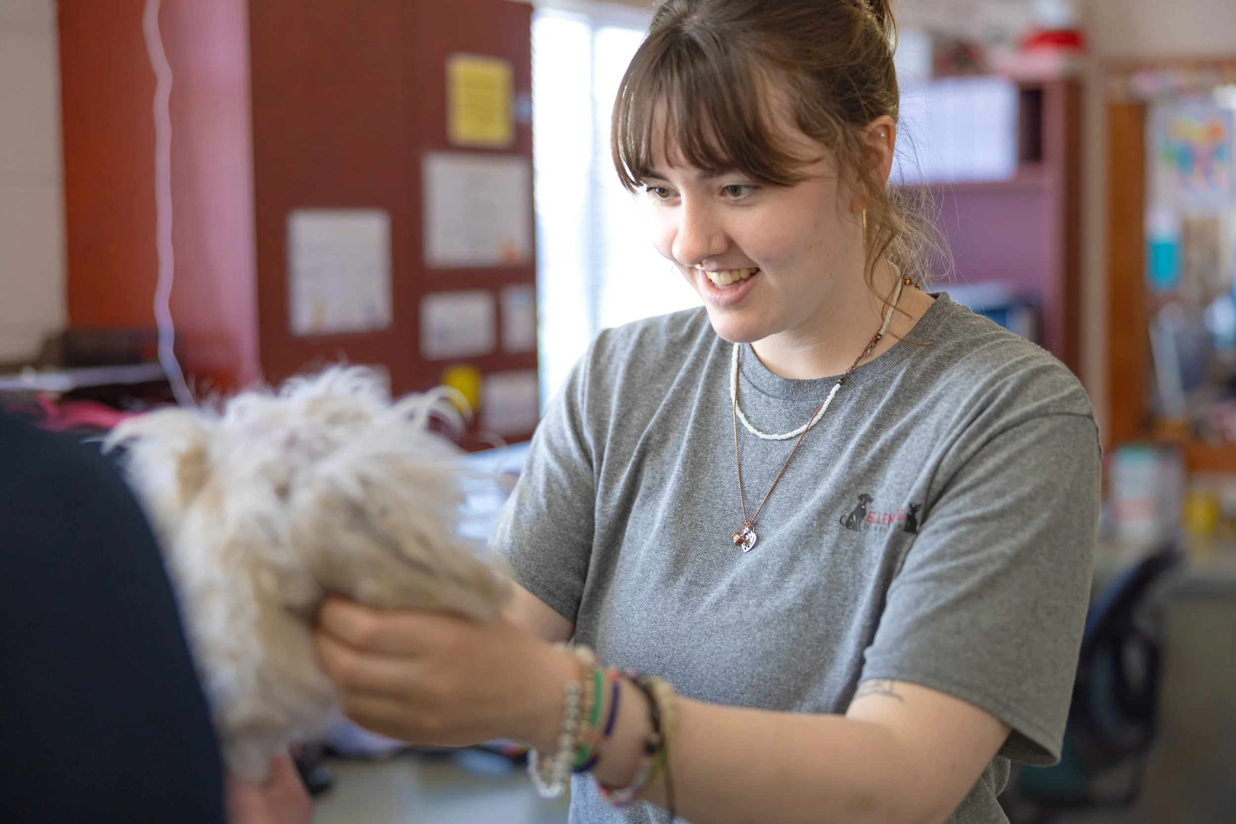 A young woman with light brown hair, wearing a gray t-shirt, multiple bracelets, and layered necklaces, is smiling and holding a small, fluffy white dog in a colorful indoor setting.