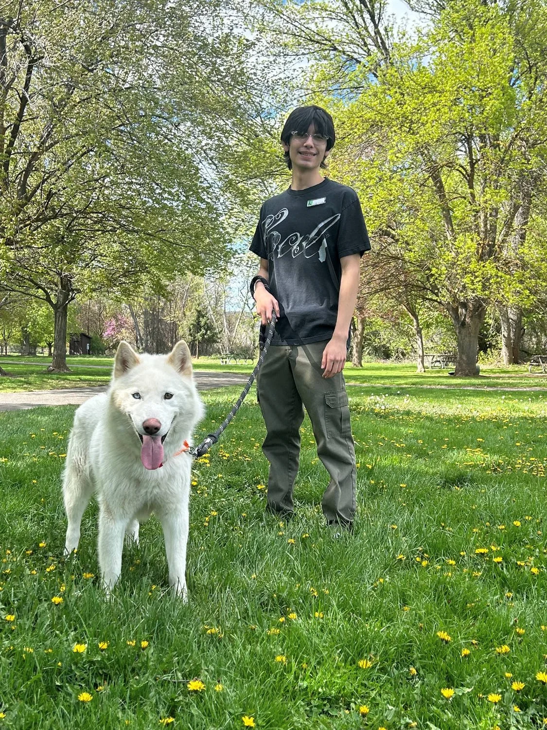 A person with black hair, glasses, wearing a black T-shirt, standing in a park with green trees and grass, holding a leash attached to a white dog.