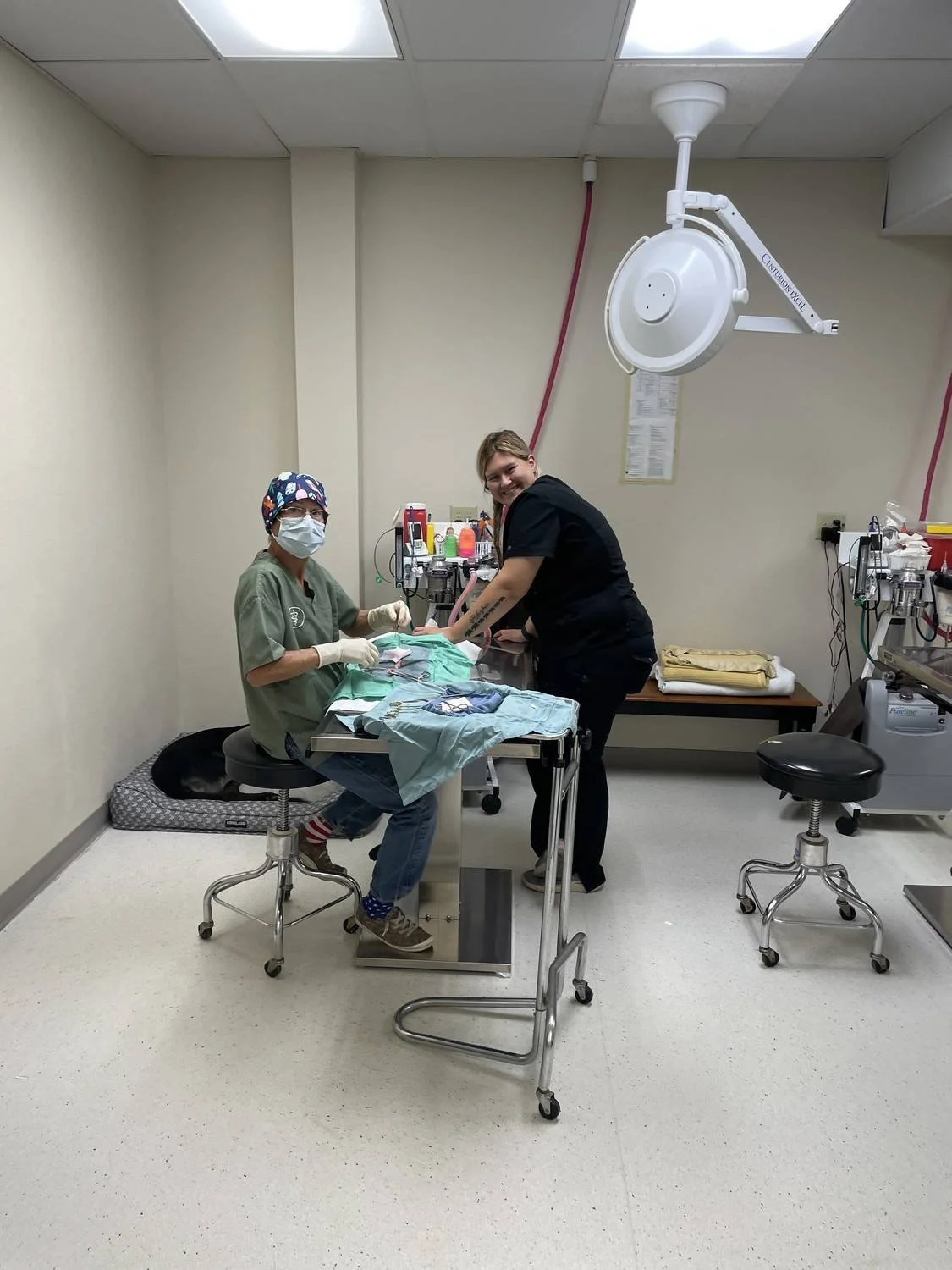 Two medical professionals preparing for a procedure in an operating room, with surgical tools and equipment on a table, one wearing a mask and scrubs, and the other smiling.