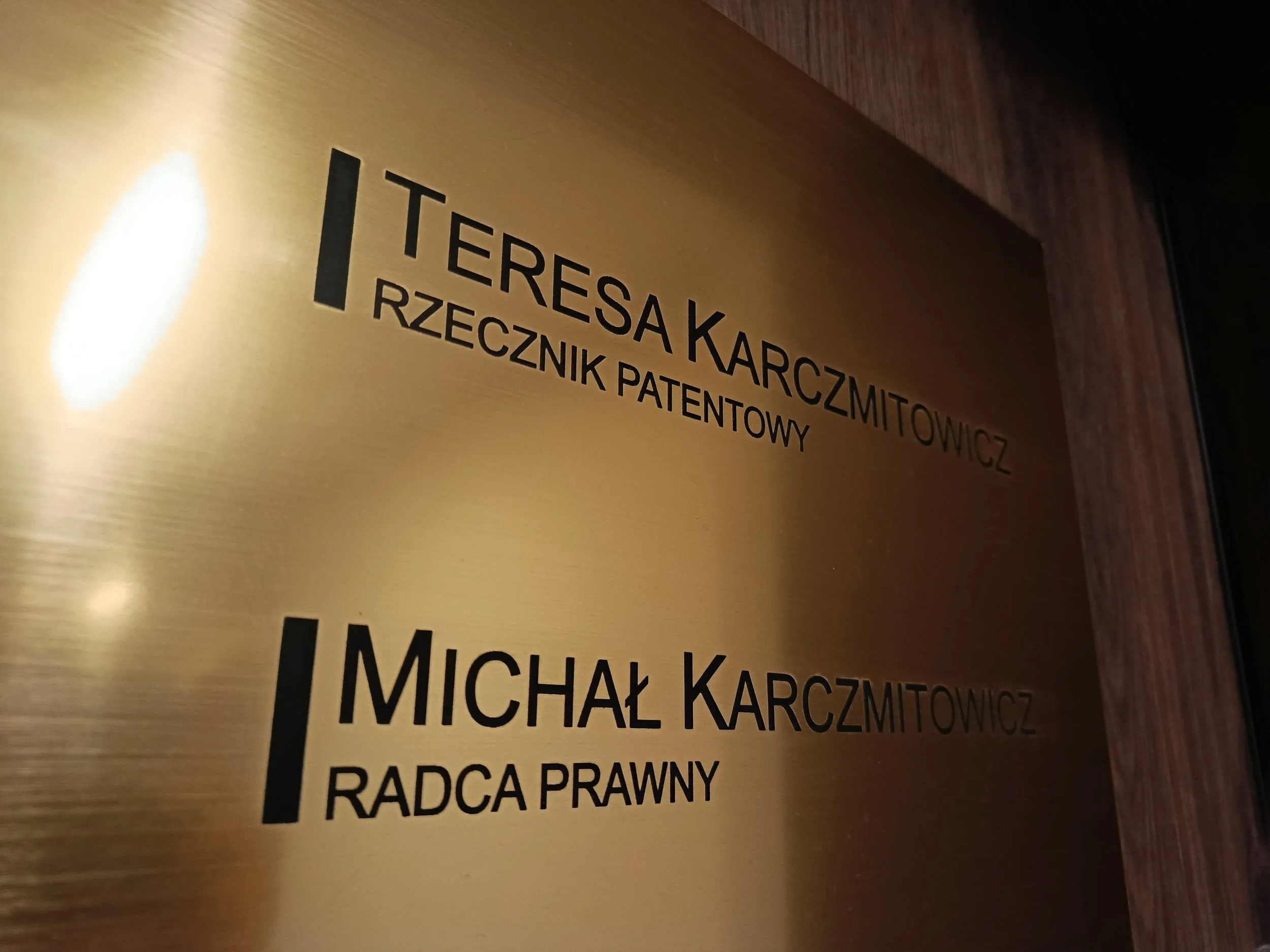 Złota tabliczka z czarnym napisem umieszczona przed drzwiami do biura, zawierająca nazwiska prawników KNM.