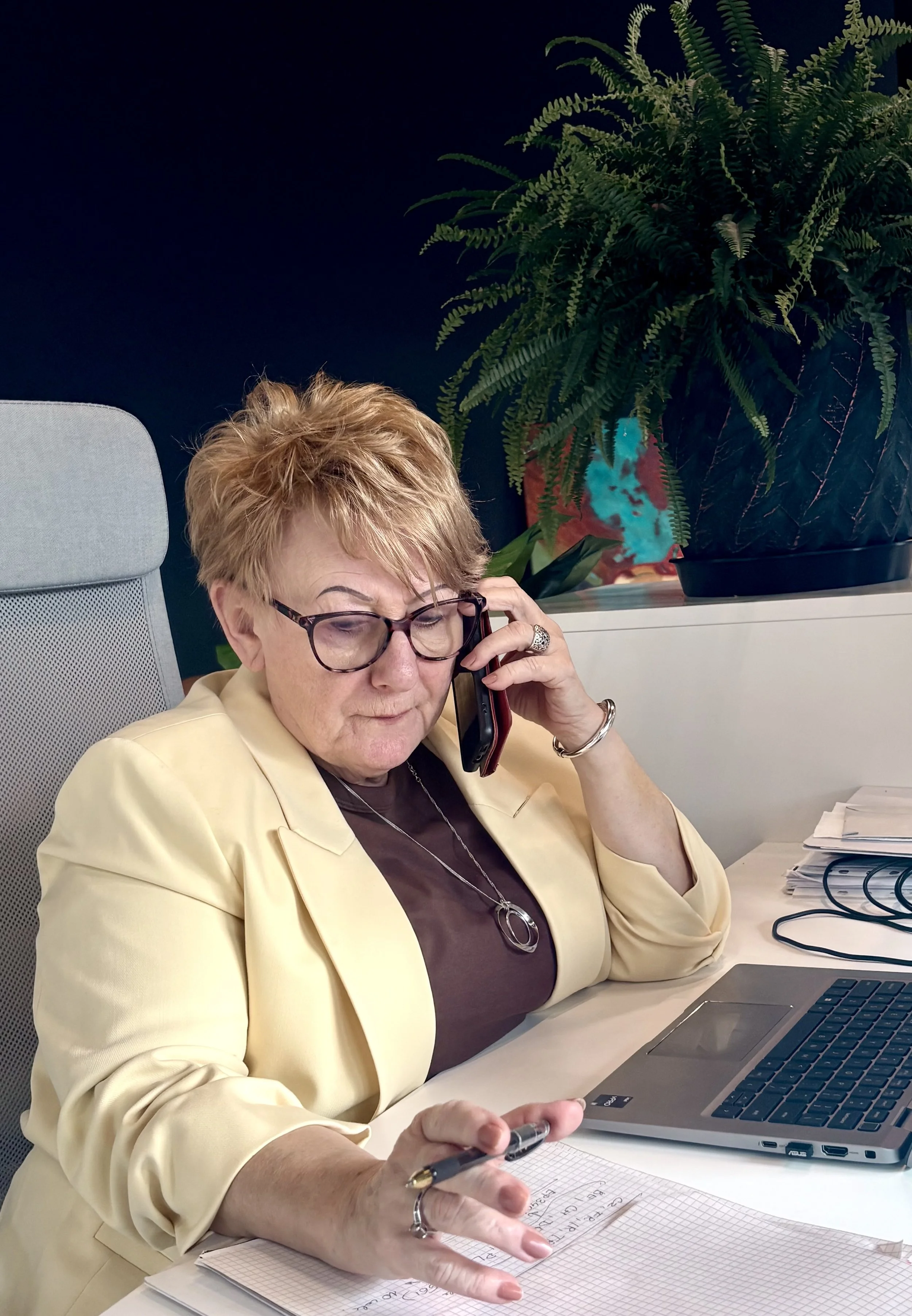 Pani Teresa dzwoni do sądu, robiąc przy tym notatki. Rozmawia przez telefon i gestykuluje lewą ręką, w której trzyma długopis.