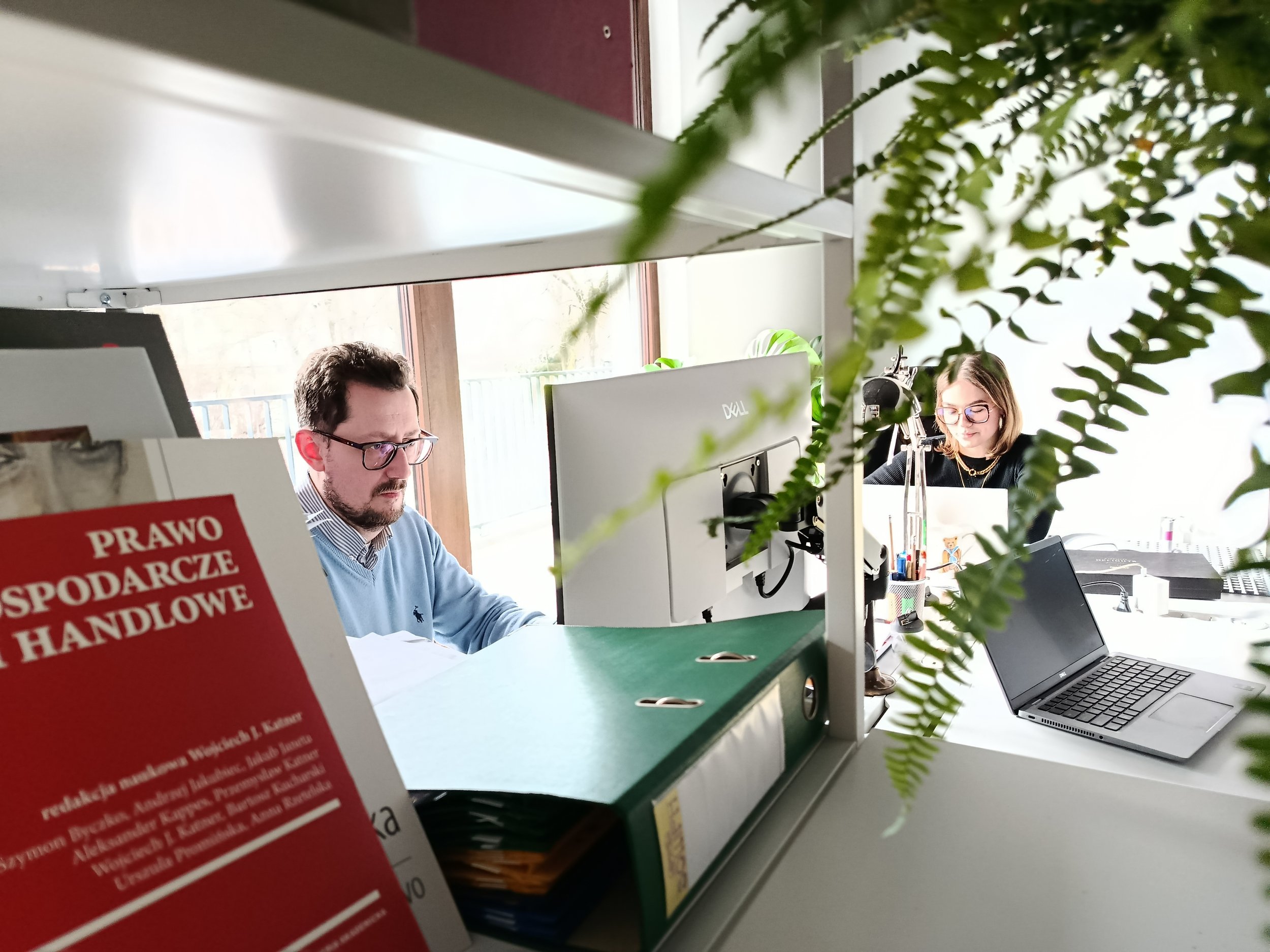 Michał i Ania pracują przy biurkach w przestrzeni biurowej KNM, częściowo zasłonięci przez zielone rośliny. Na pierwszym planie widać książkę poświęconą prawu gospodarczemu i handlowym.