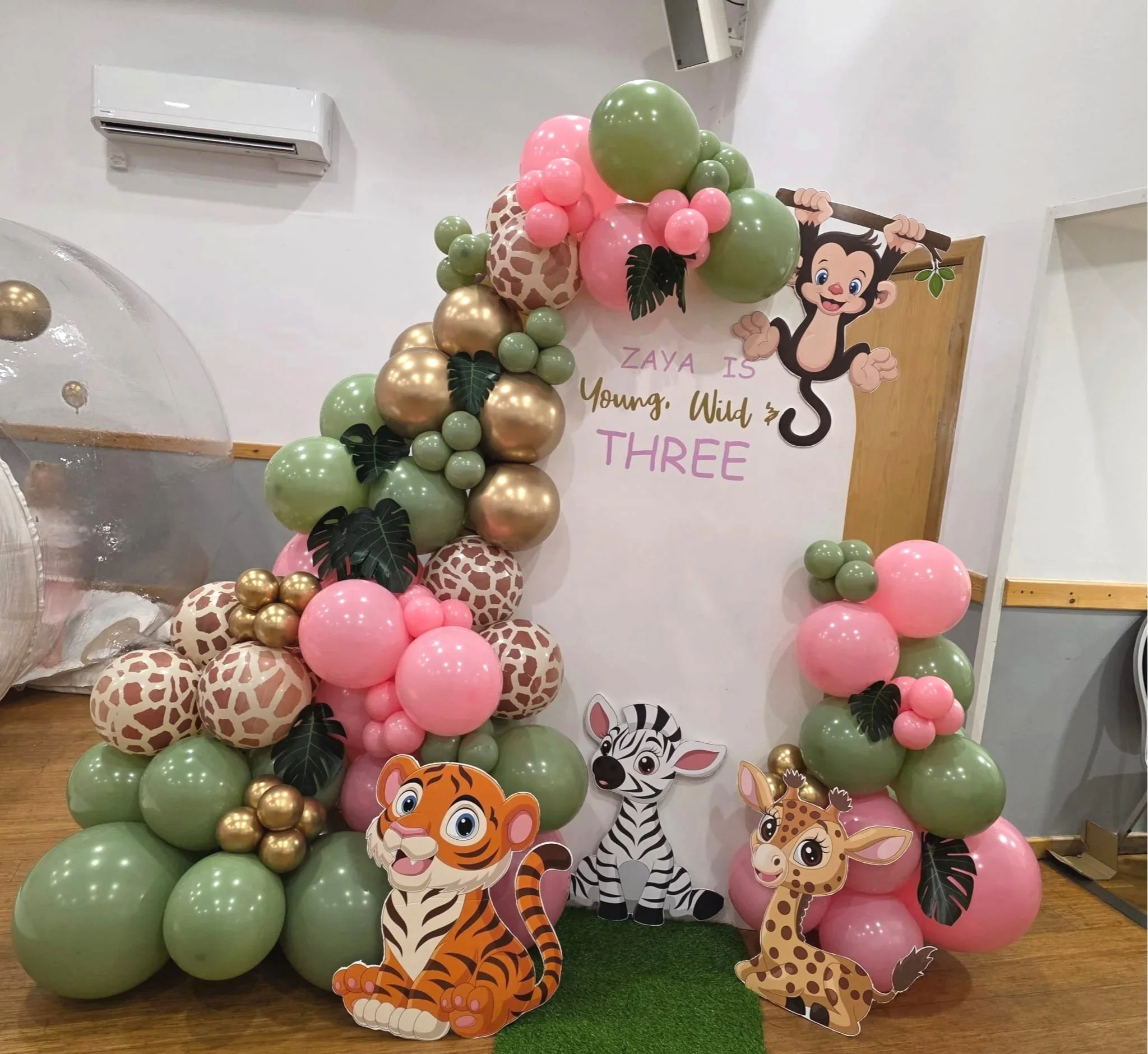 Decorative jungle-themed birthday backdrop with balloons, cartoon animal cutouts, and a message that says 'Zaya is Young, Wild & Three'.