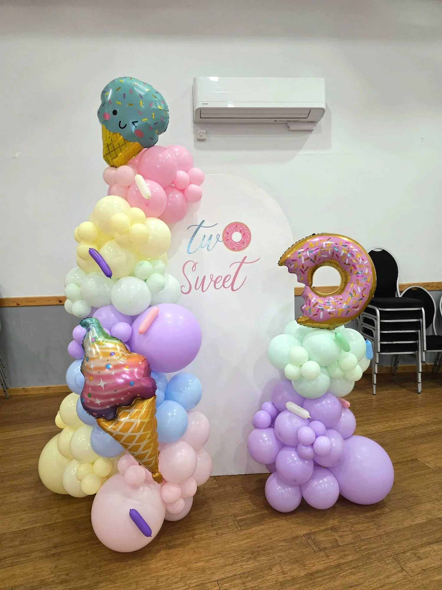 Colorful balloon decorations with ice cream cone and donut shaped balloons, and a white backdrop that reads 'two sweet', set in a room with wooden floor and black chairs.