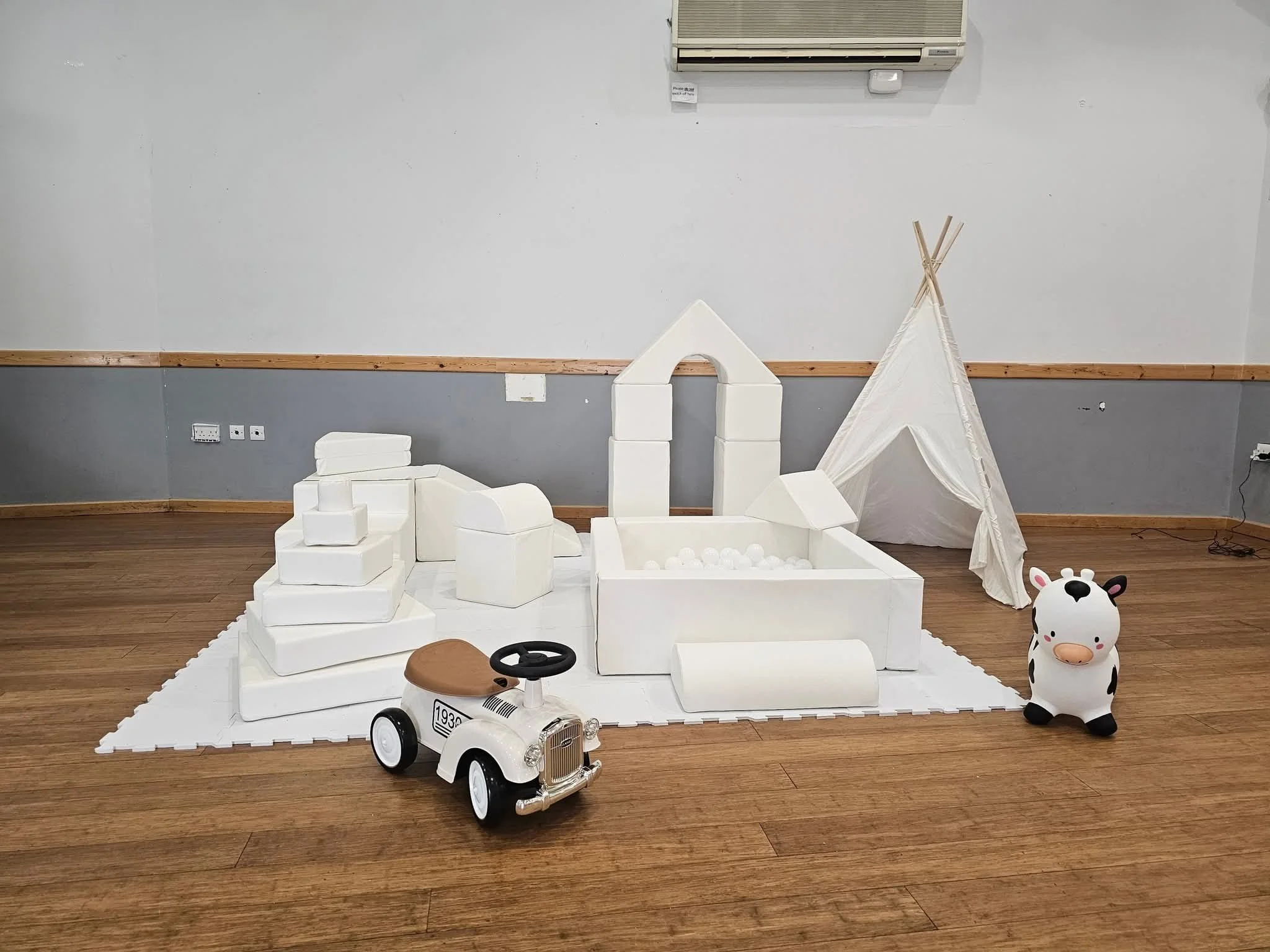Children's soft play hire with white foam blocks, a tipi tent, a toy car, and a cow-shaped ride-on toy on a white foam mat on a wooden floor.