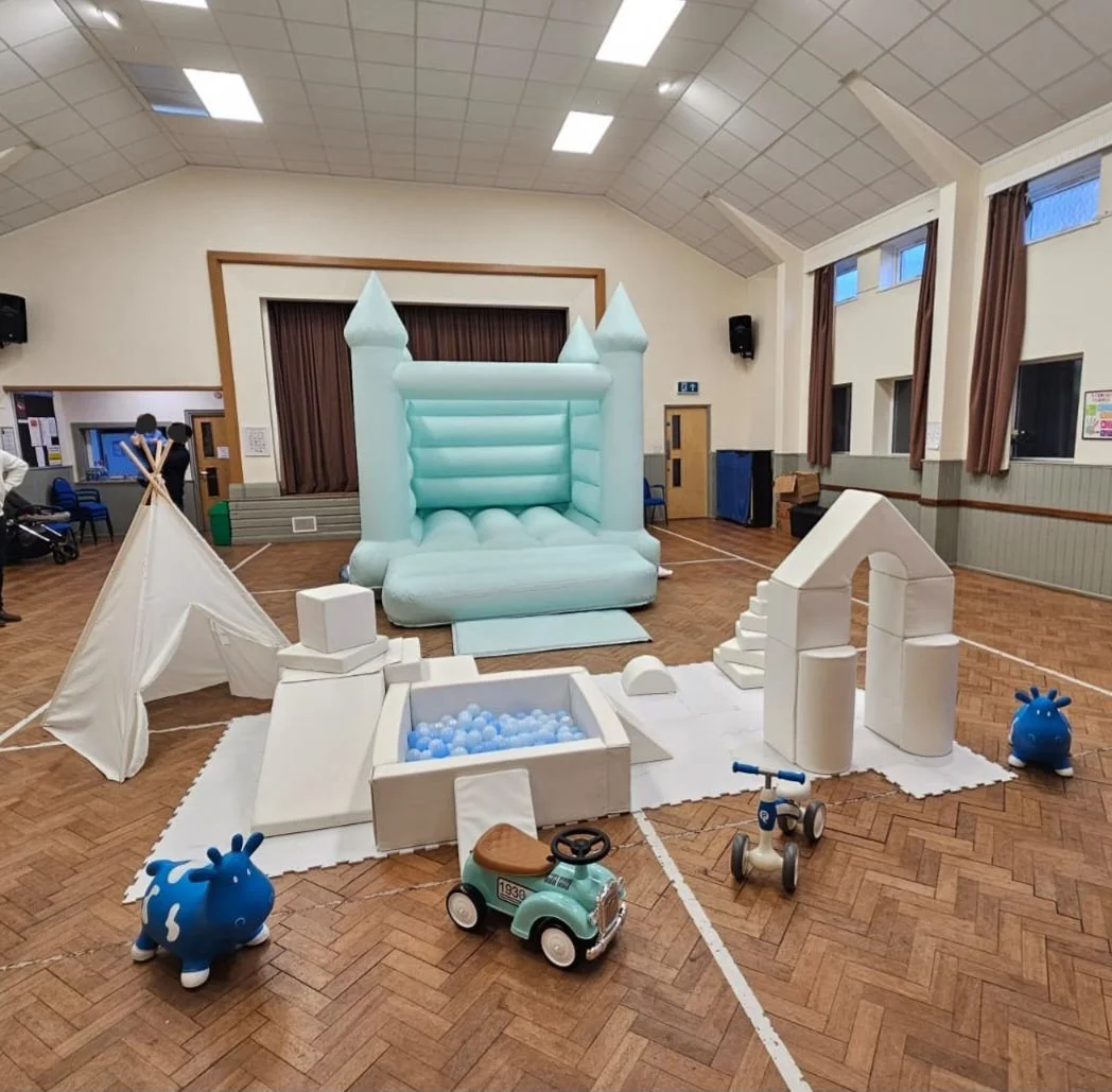 Indoor soft play hire with a blue bouncy castle, small tent, ball pit, archway, toy cars, and plush animal toys in a community hall or gymnasium.