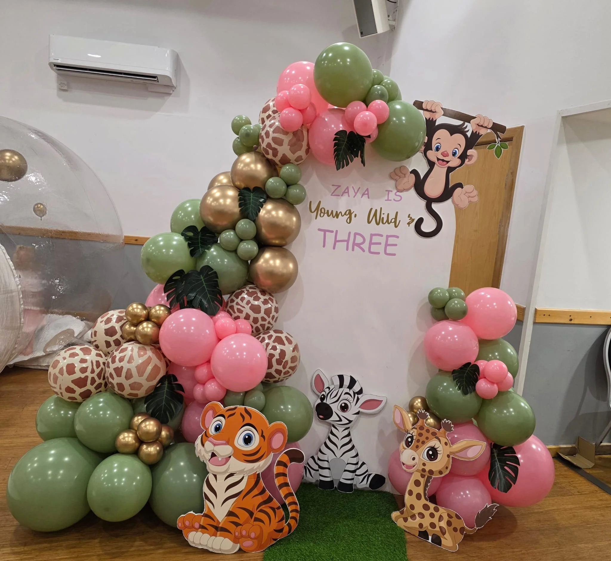 Decorative jungle-themed birthday backdrop with balloons, cartoon animal cutouts, and a message that says 'Zaya is Young, Wild & Three'.