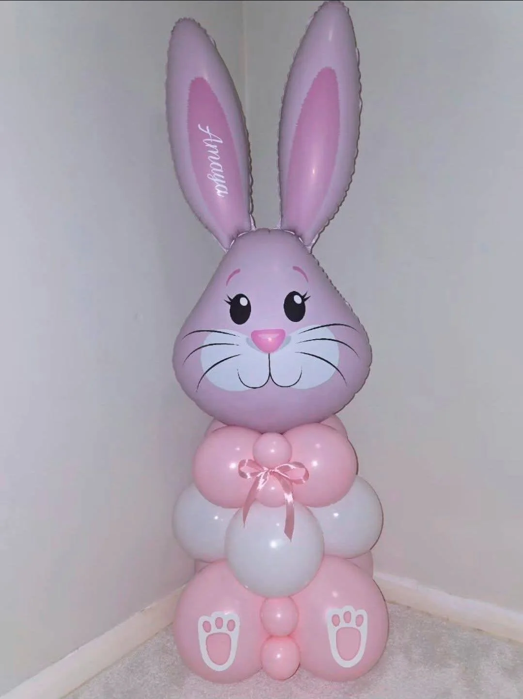 Pink bunny-shaped balloon stack with tall ears, a smiling face, and a bow on its chest, standing on a beige carpeted floor against a plain wall.