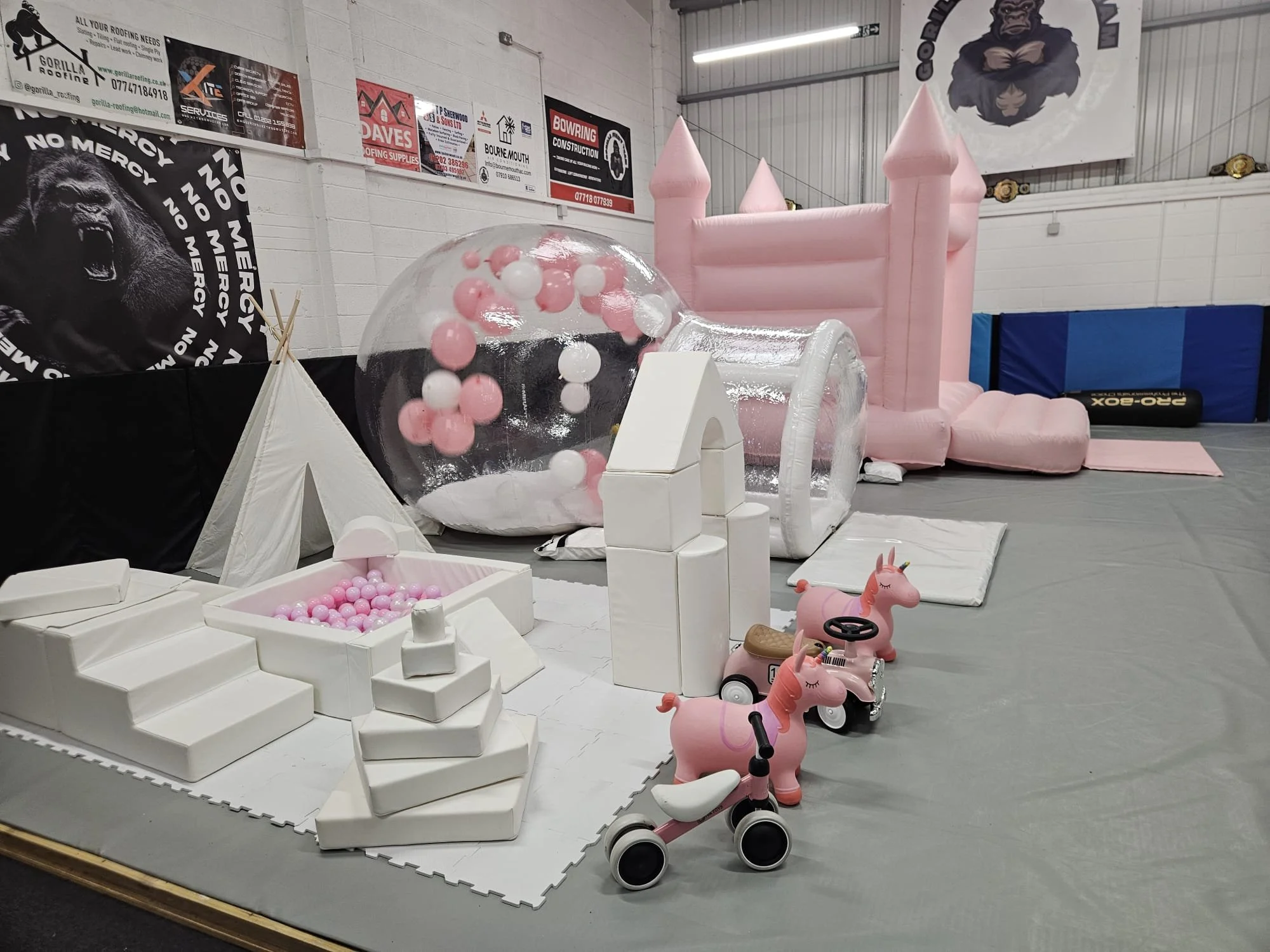 Indoor play area with pink bouncy inflatable castle, small pink ride-on unicorn toys, a pink ball pit, and a bubble house with pink and white balls.