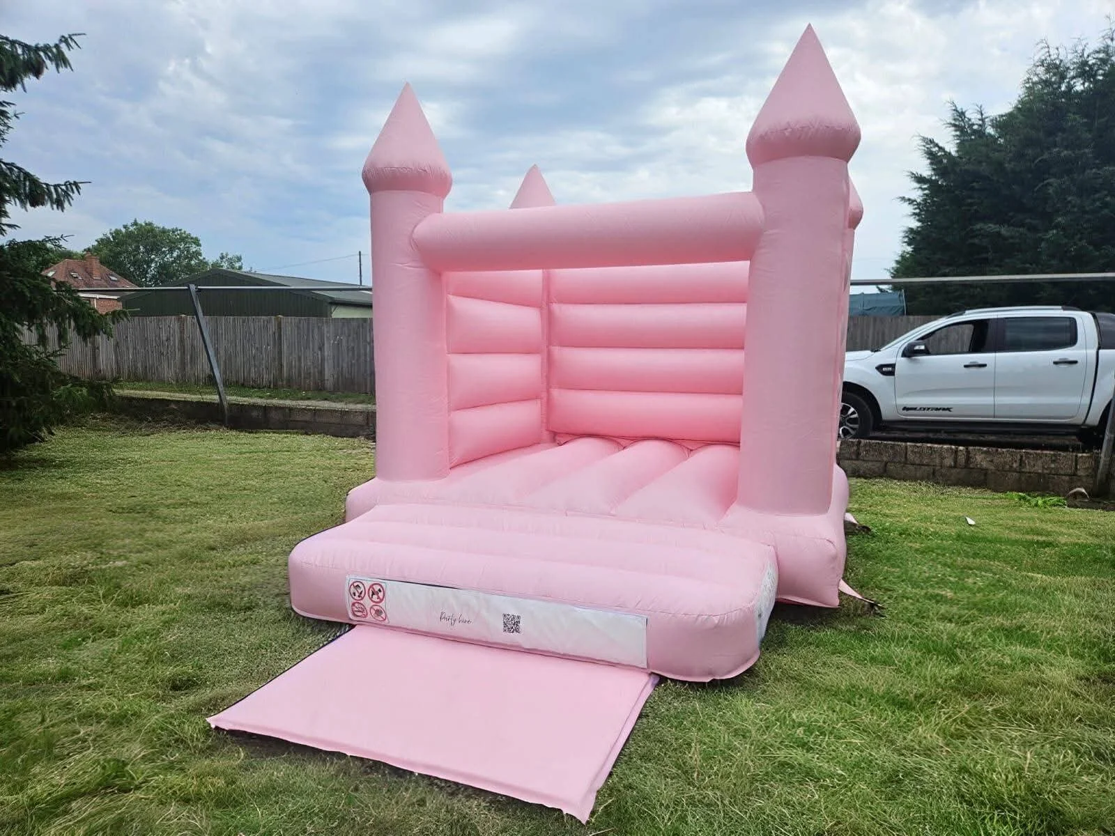 A pink bouncy castle with a small slide outside on a grassy lawn, situated in a backyard with a wooden fence, trees, a house, and a white pickup truck in the background.