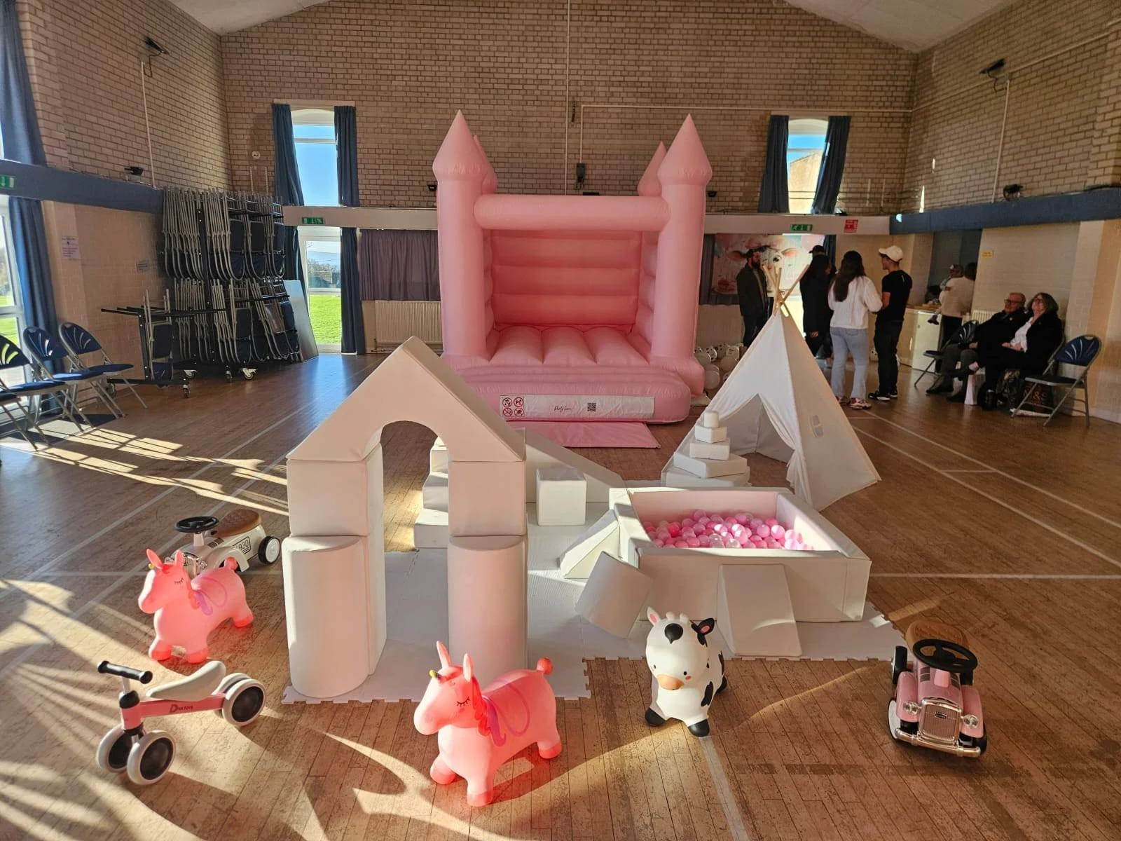 Indoor children's party with pink bouncy castle, white tents, pink and white toy unicorns, a small pink toy car, and a ball pit filled with pink and white balls. In the background, there are people standing and sitting, with windows and brick walls.