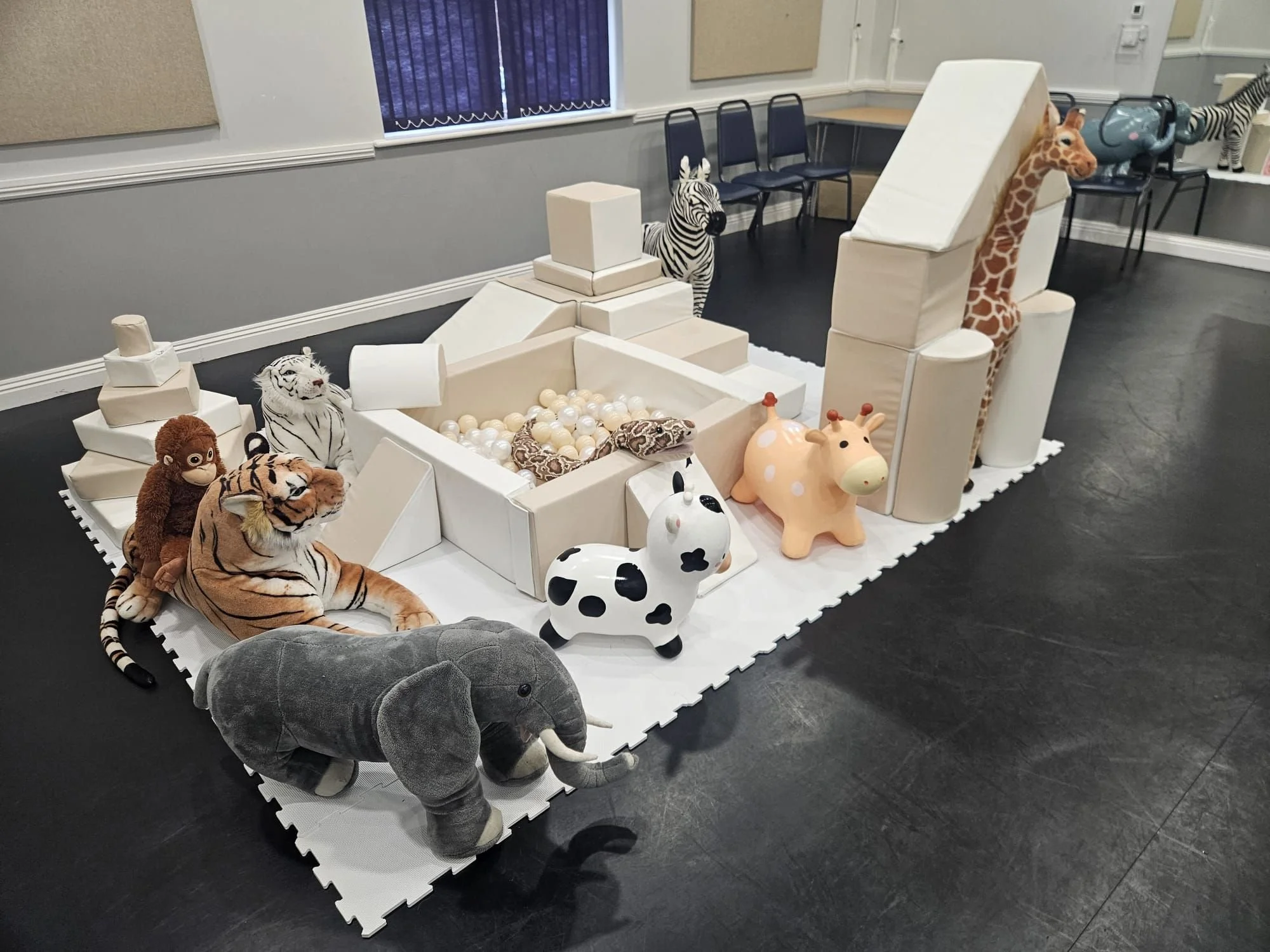Soft play hire with stuffed animal toys resembling a jungle, including a lion, tiger, zebra, giraffe, cow, pig, elephant, and monkey, arranged on a foam puzzle mat with various foam structures and a ball pit.