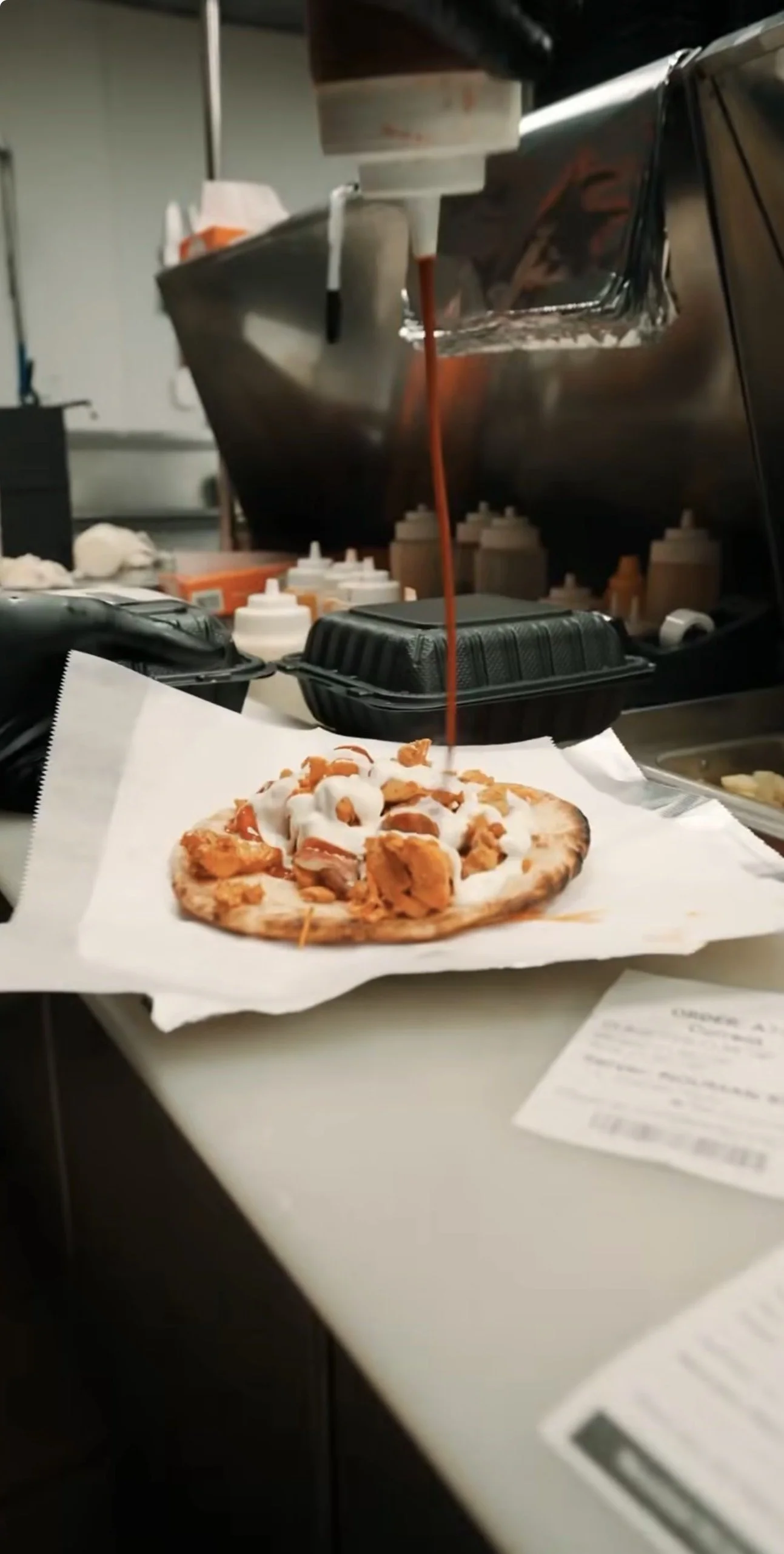 A pizza topped with fried chicken pieces, white sauce, and tomatoes with sauce being poured over it.