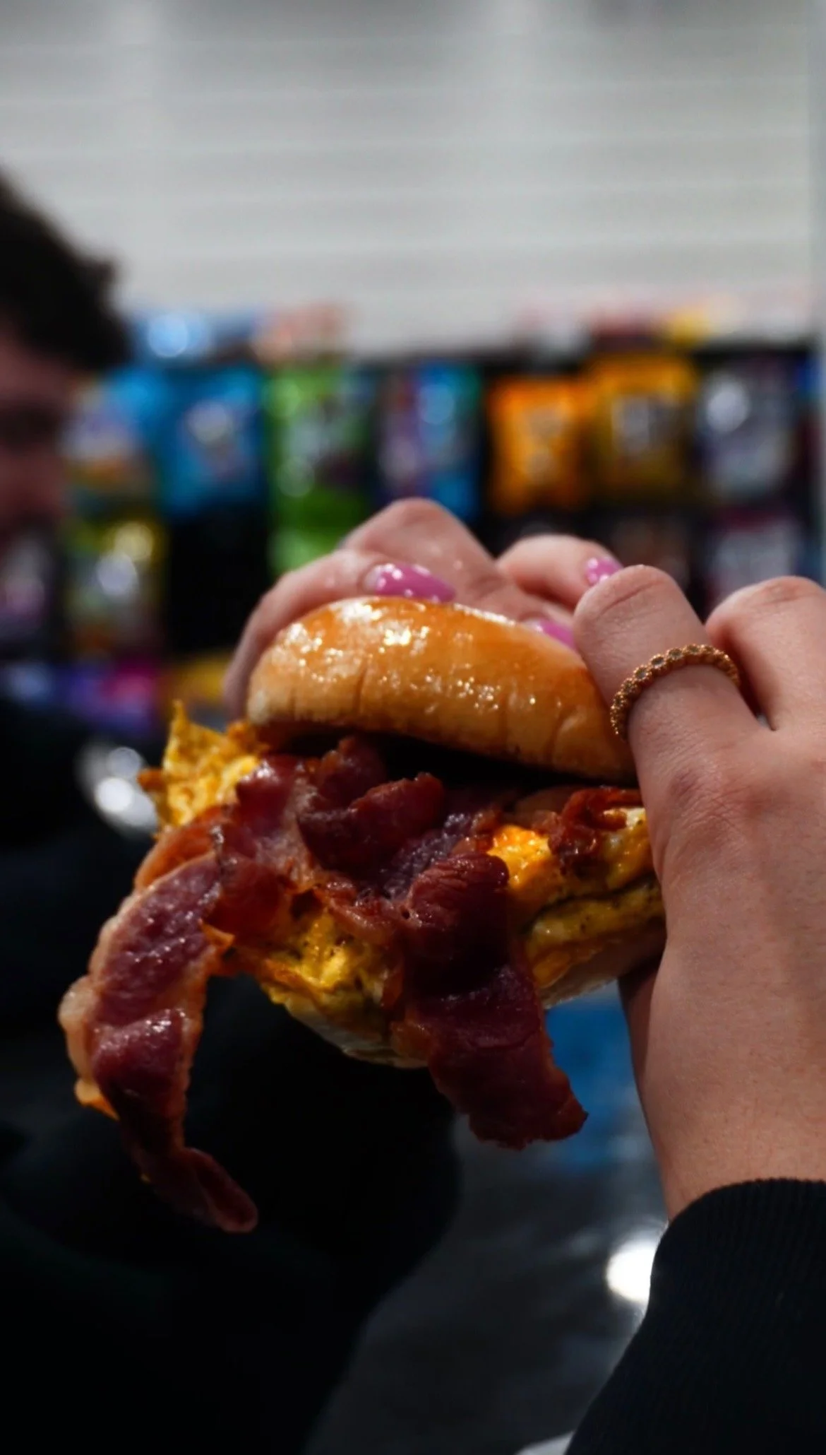 A person holding a bacon, egg, and cheese breakfast sandwich with a bun, eggs, and bacon in a store or supermarket aisle.