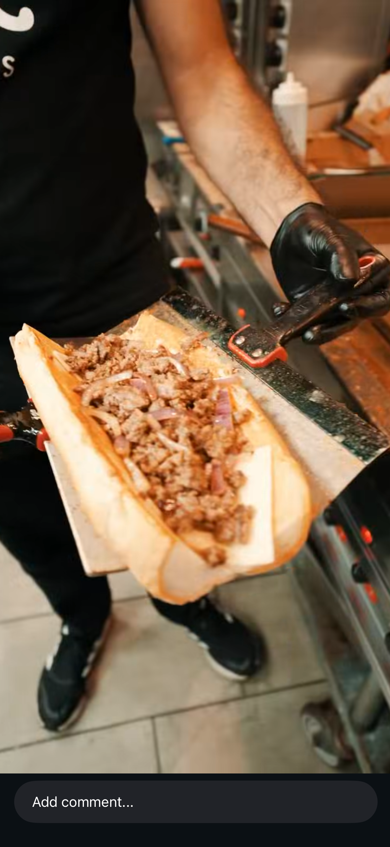 Person wearing black gloves holding a sandwich bun filled with cooked ground meat and sliced onions, likely in a commercial kitchen or restaurant setting.