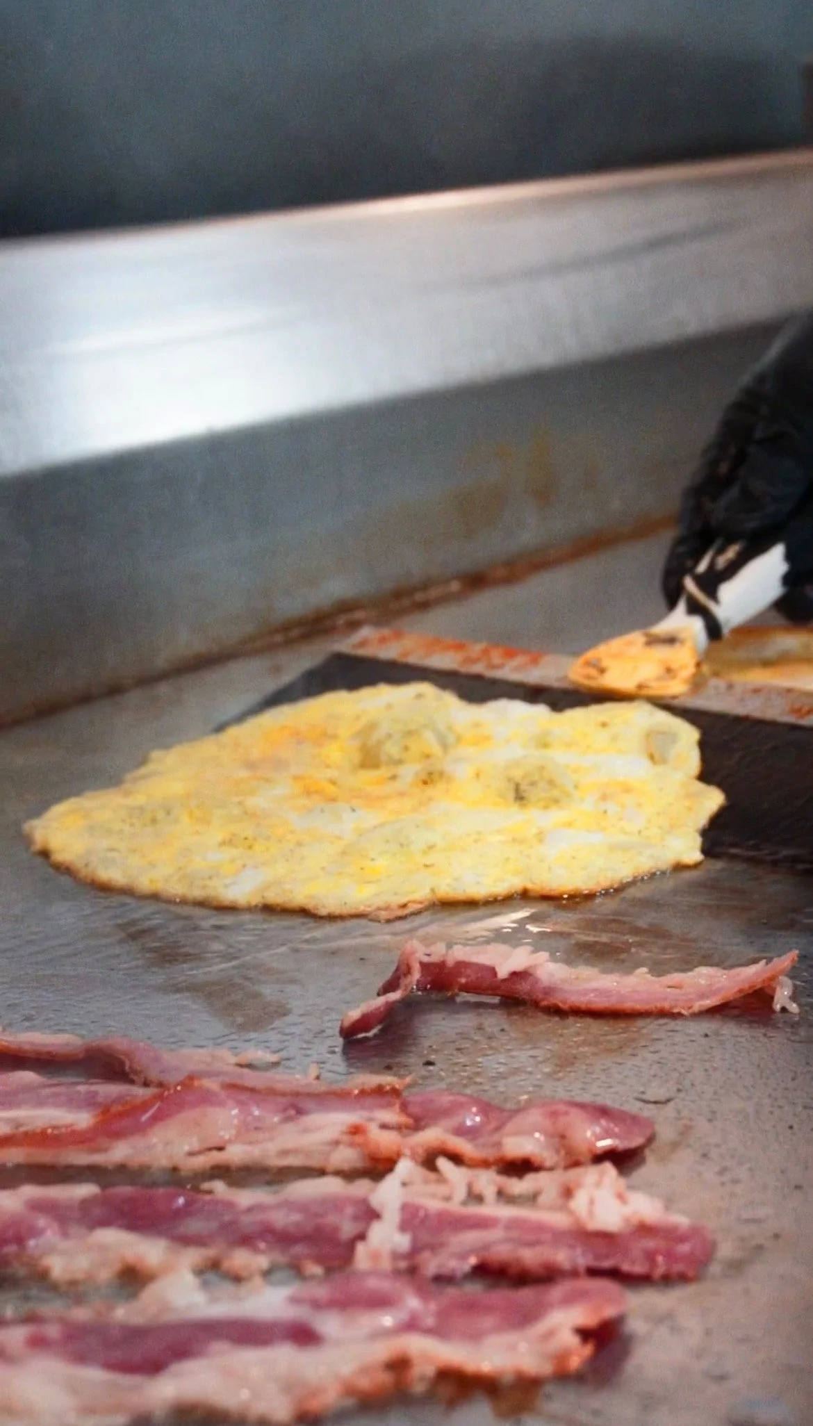 Eggs cooking on a griddle with bacon strips in the foreground.