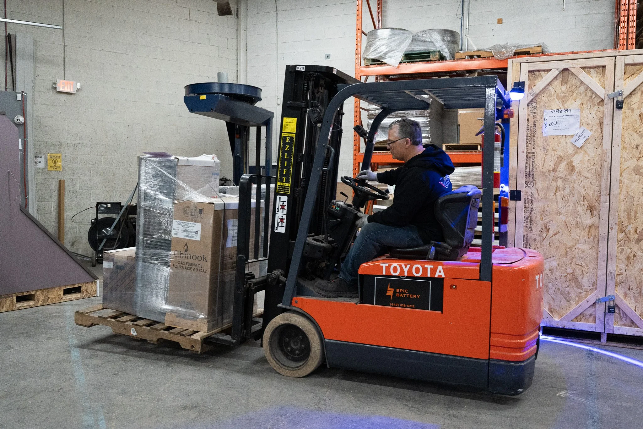 A man driving a Toyota forklift truck inside a warehouse, moving a pallet with boxes wrapped in plastic.