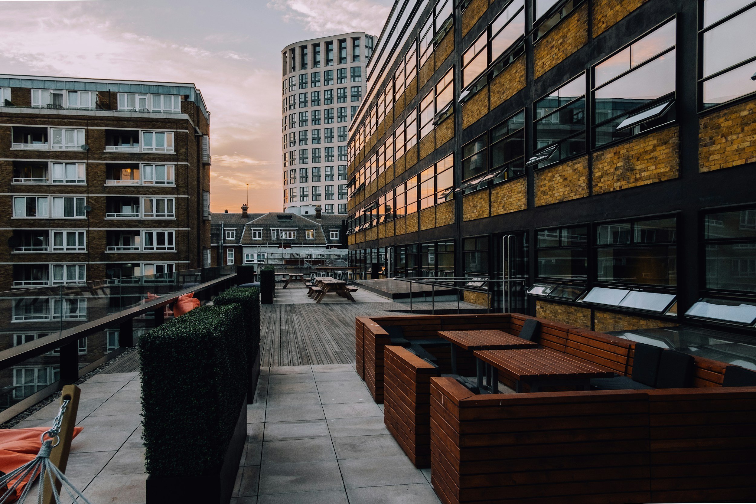 A city rooftop terrace at sunset with seating areas, wooden benches, a table, and surrounding buildings with reflective windows.