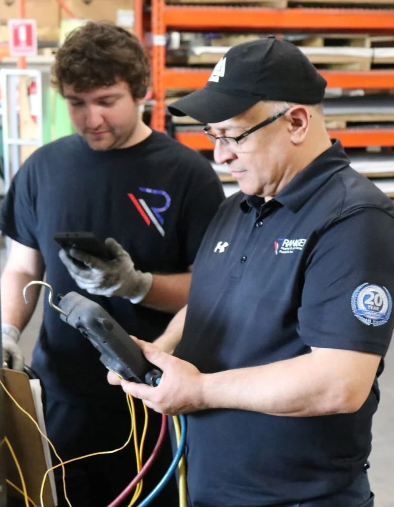 Two men working in a warehouse or store with shelves of materials; one is holding a diagnostic device with wires, the other is using a smartphone, both examining equipment.