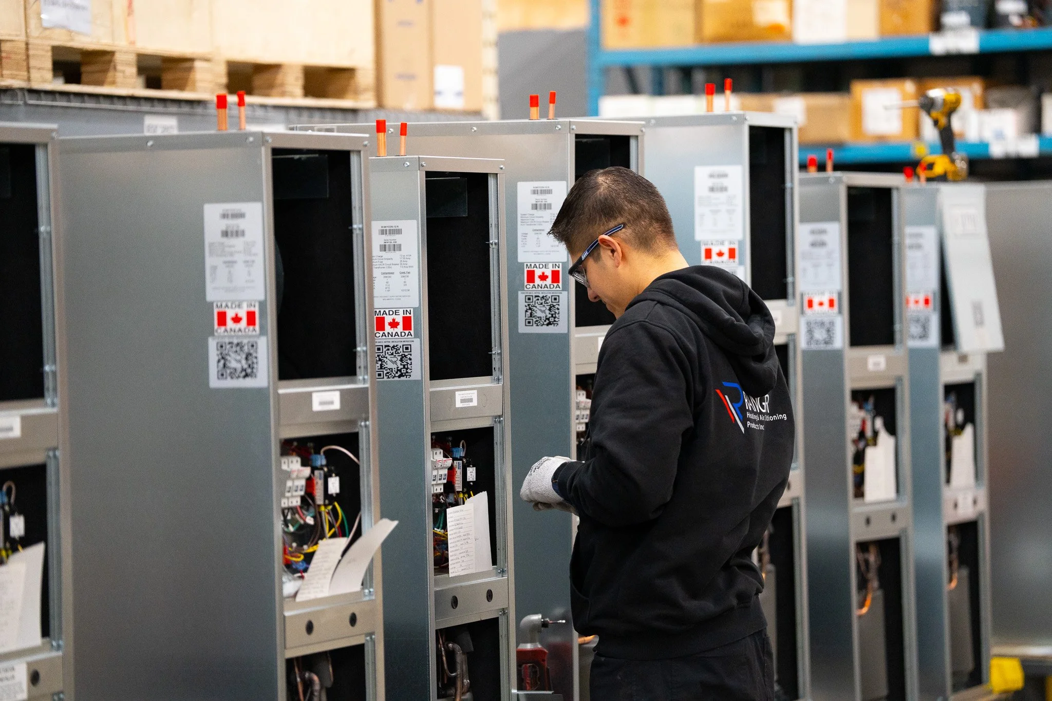 A man working on electrical panels in a warehouse, wearing a black hoodie with company logo, safety gloves, and safety glasses.