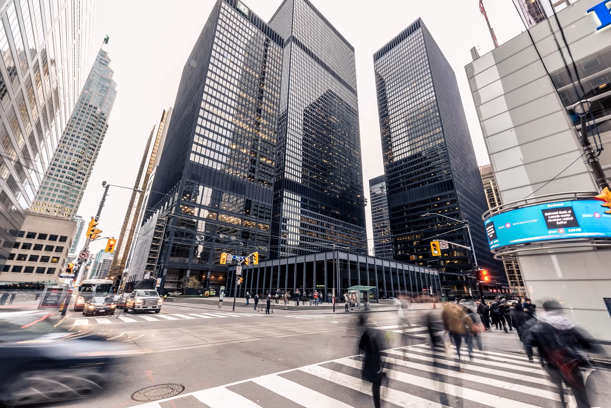 Tall reflective skyscrapers in a busy city intersection with people crossing on a crosswalk, cars, and traffic lights.