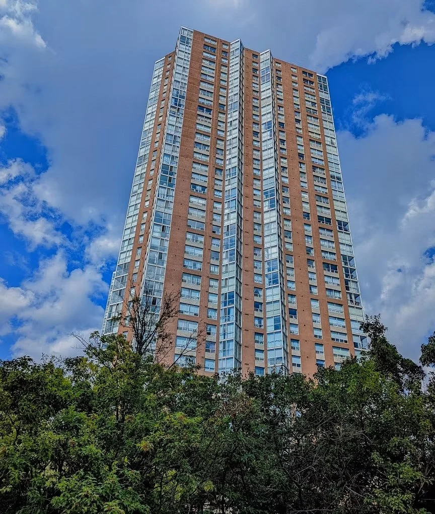 A tall residential skyscraper with many windows, viewed from the ground with trees at the base, against a partly cloudy blue sky.