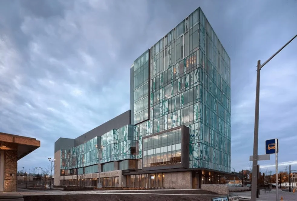 Modern multi-story building with a glass facade reflecting the cloudy sky, located on a city street with street signs and lamps.