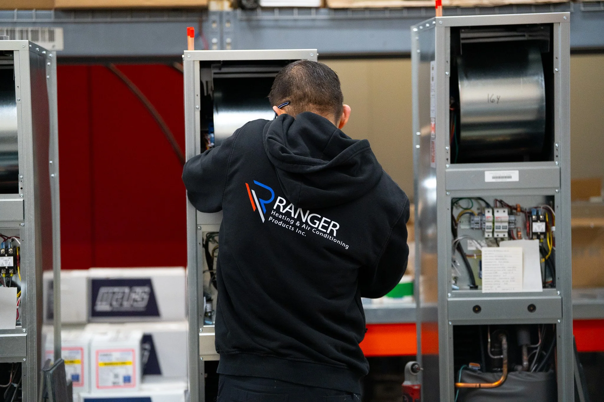 A technician working on HVAC or refrigeration equipment in a warehouse, wearing a black hoodie with a company logo that reads 'RANGER Heating & Air Conditioning Products Inc.'