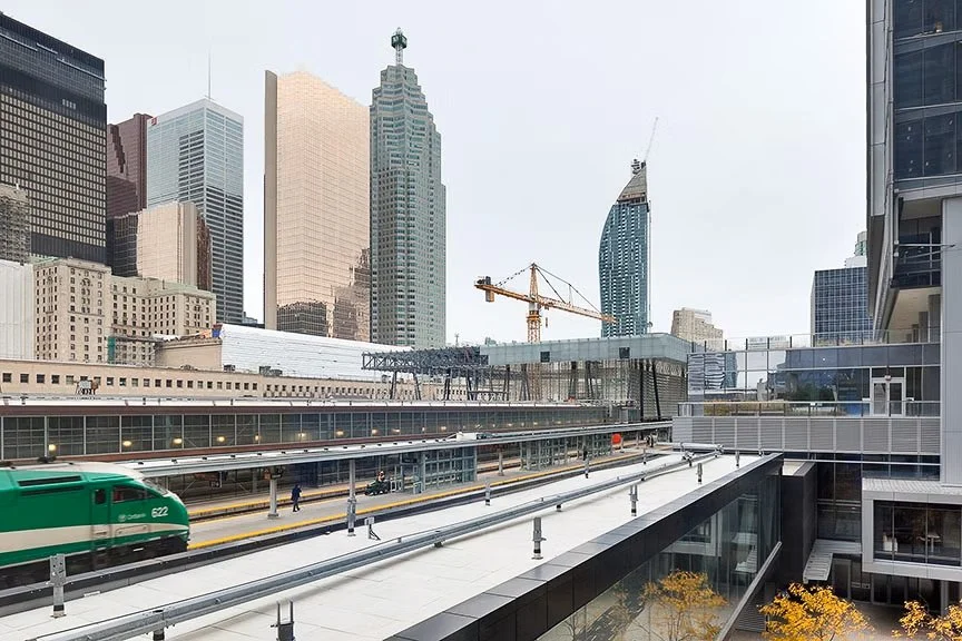Cityscape with tall skyscrapers, a green train moving on elevated tracks, and construction cranes in the background, viewed from a modern building''s balcony.